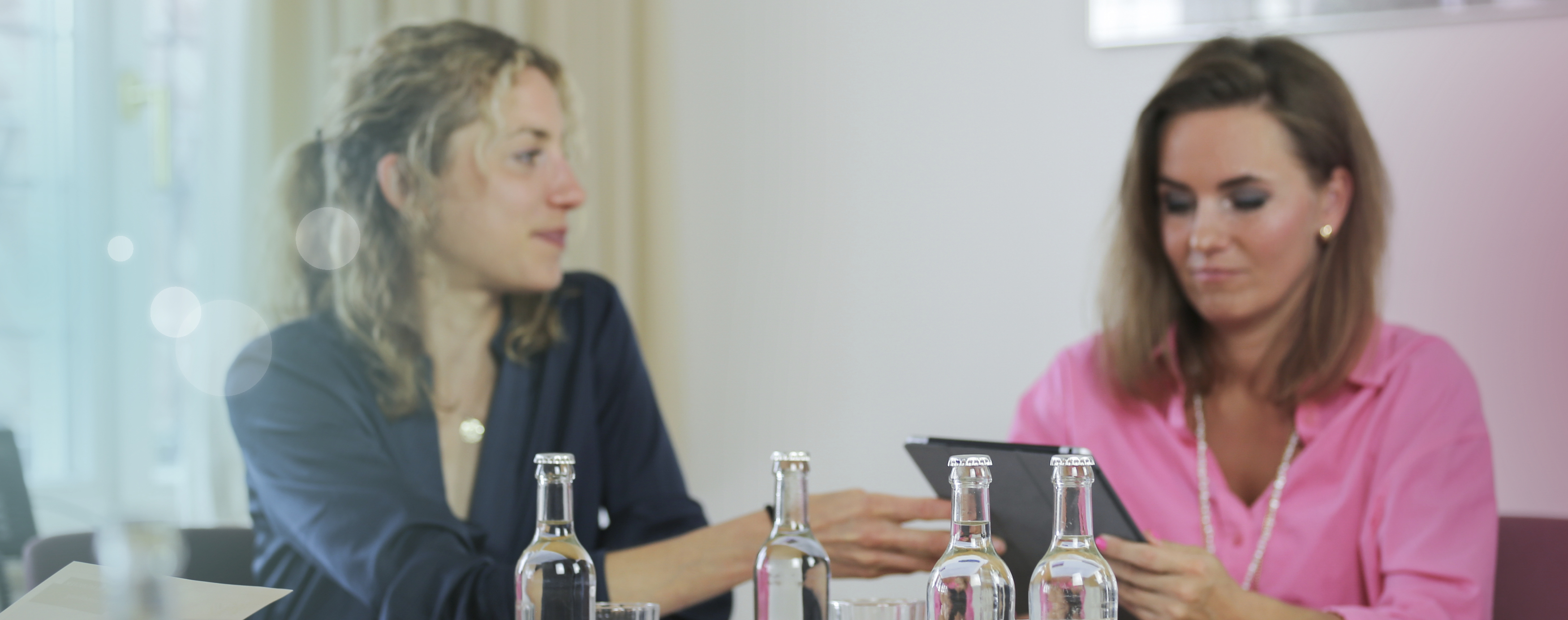 Zwei junge Frauen sitzen gemeinsam an einem Tisch und besprechen etwas auf einem Tablet, im Vordergrund zeichnen sich Wasserflaschen aus Glas ab.