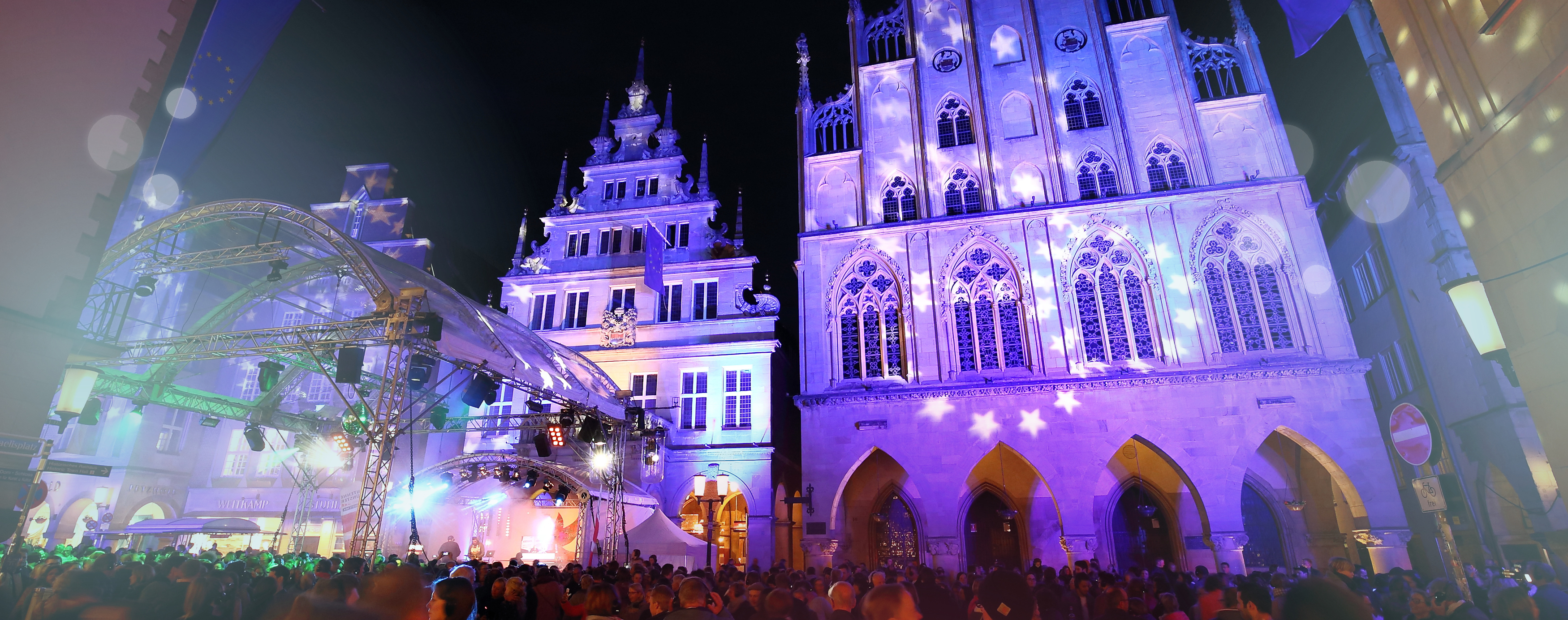 Bunt beleuchtet, bei Nacht, zu sehen sind Häuser des Prinzipalmarkts, vor ihnen eine Bühne, um die sich viele Menschen versammeln und feiern.