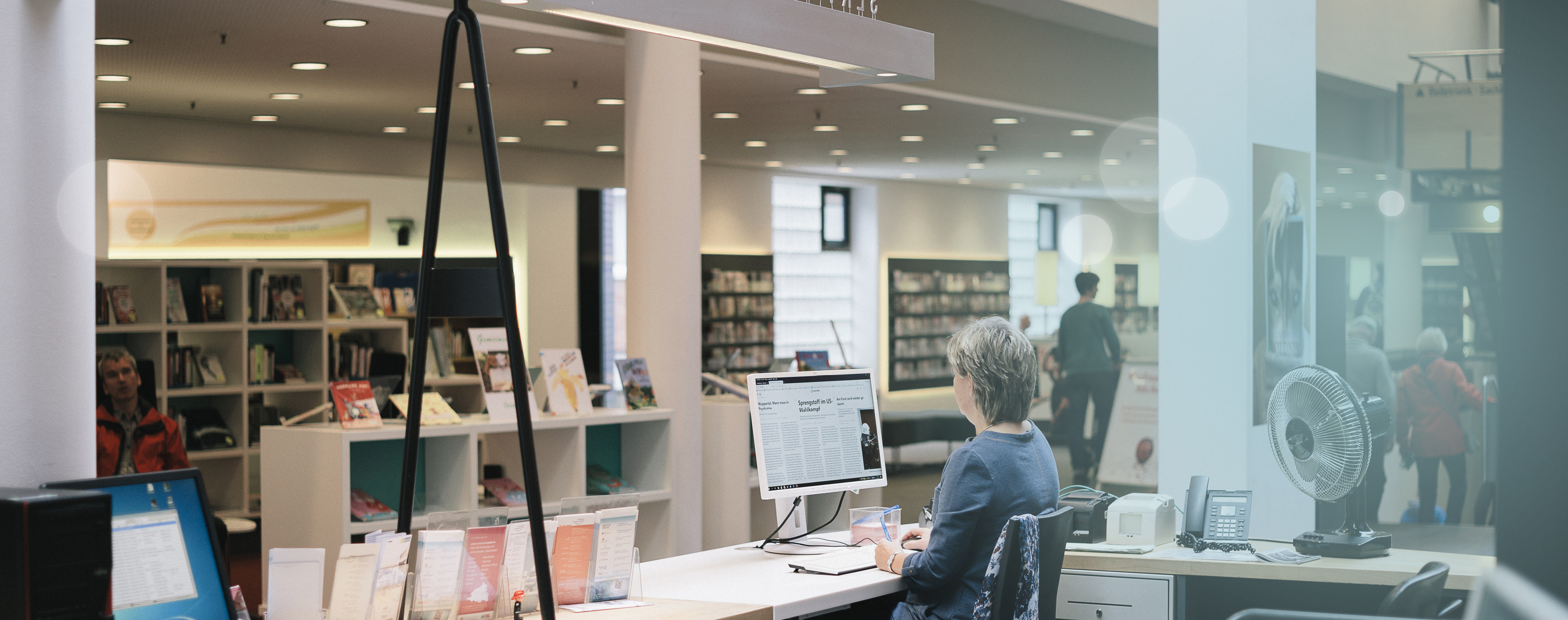 Eine Frau sitzt an einem Computer,  im Hintergrund sieht man Menschen und viel volle Bücherregale.