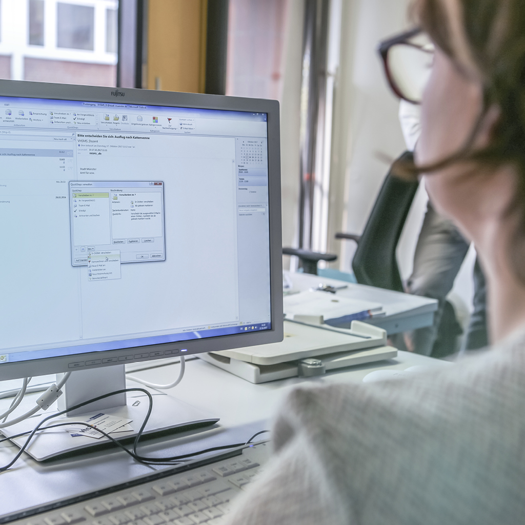 Eine im Vordergrund unscharf fotografierte Frau mit Brille arbeitet an einem Computer, der scharf im Hintergrund zu erkennen ist.