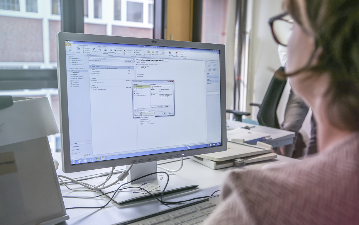 Eine im Vordergrund unscharf fotografierte Frau mit Brille arbeitet an einem Computer, der scharf im Hintergrund zu erkennen ist.