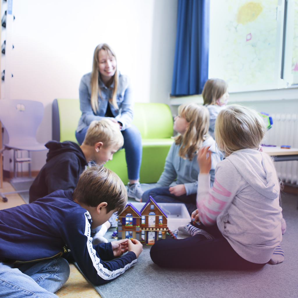 Fünf Grundschulkinder spielen und unterhalten sich im Vordergrund, die Erzieherin sitzt auf dem Sofa dahinter und beobachtet die Situation.