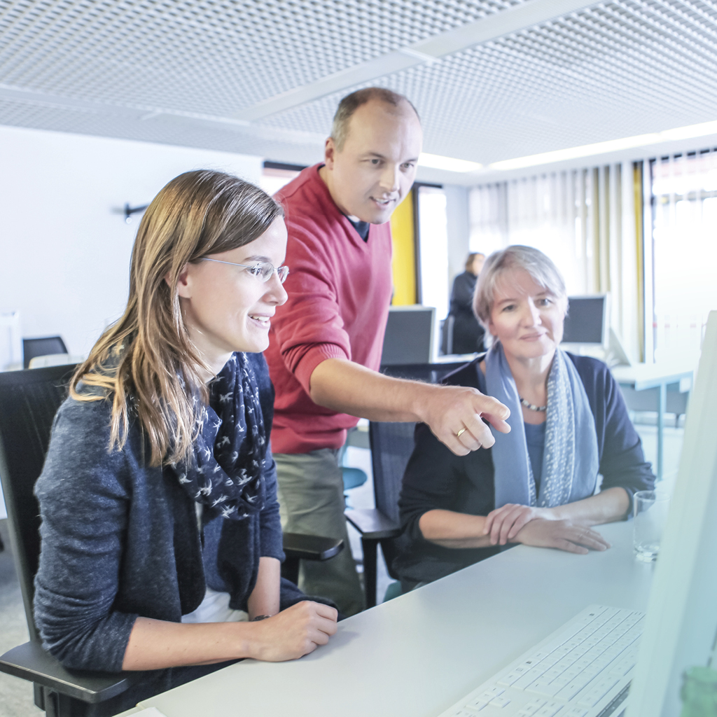 Zwei am Tisch sitzende Frauen schauen freundlich auf einen Computerbildschirm, der dahinter stehende Mann zeigt mit seinem Arm auf etwas auf dem Bildschirm.