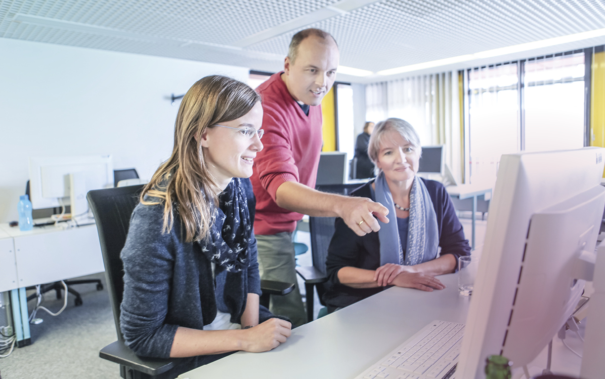 Zwei am Tisch sitzende Frauen schauen freundlich auf einen Computerbildschirm, der dahinter stehende Mann zeigt mit seinem Arm auf etwas auf dem Bildschirm.