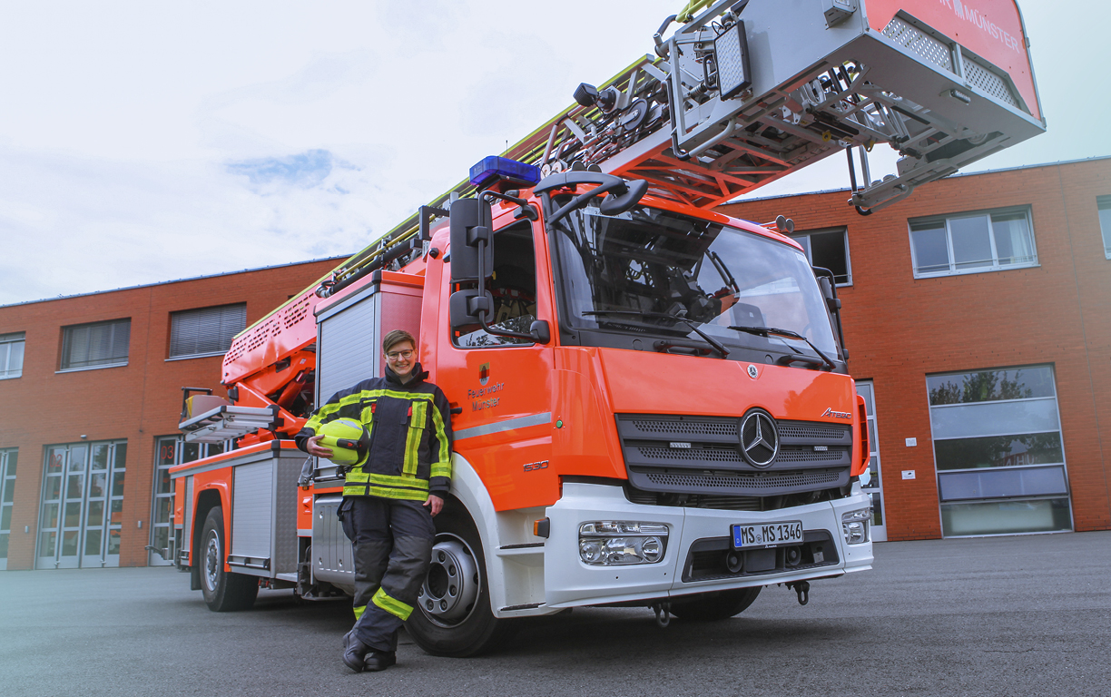 Eine weibliche Feuerwehr-Fachkraft steht vor einem Drehwagen, der vor einer Feuerwache steht, uns lächelt in die Kamera.