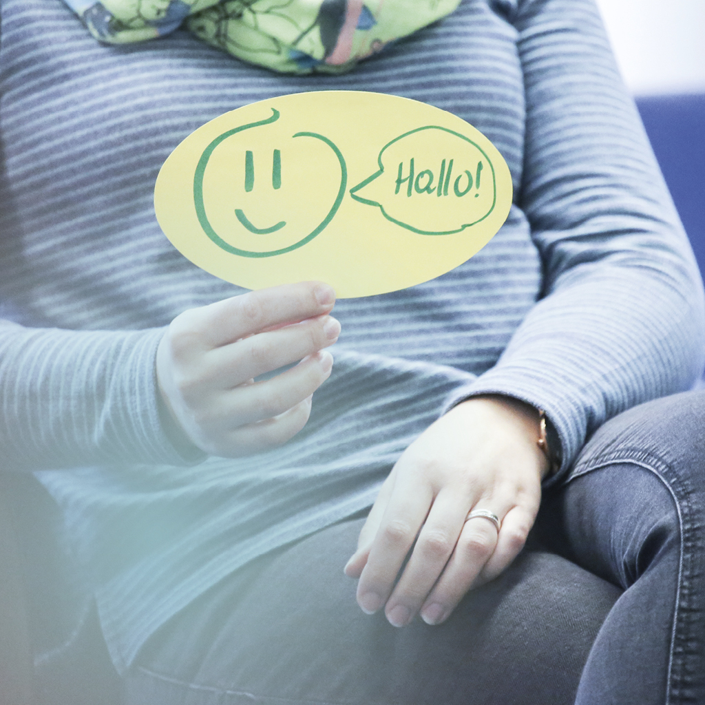 Eine sitzende Frau hält ein Karton in der Hand, auf dem ein Smiley mit einem Hallo zu sehen ist.