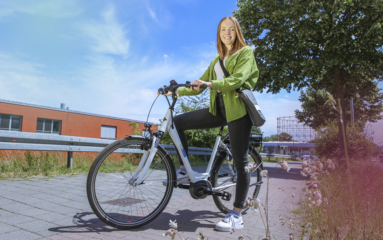 Eine rothaarige Frau trägt ein hellgrünes Hemd und sitzt auf einem Dienst-E-Bike, welches mit dem Logo der Stadt Münster bedruckt ist.