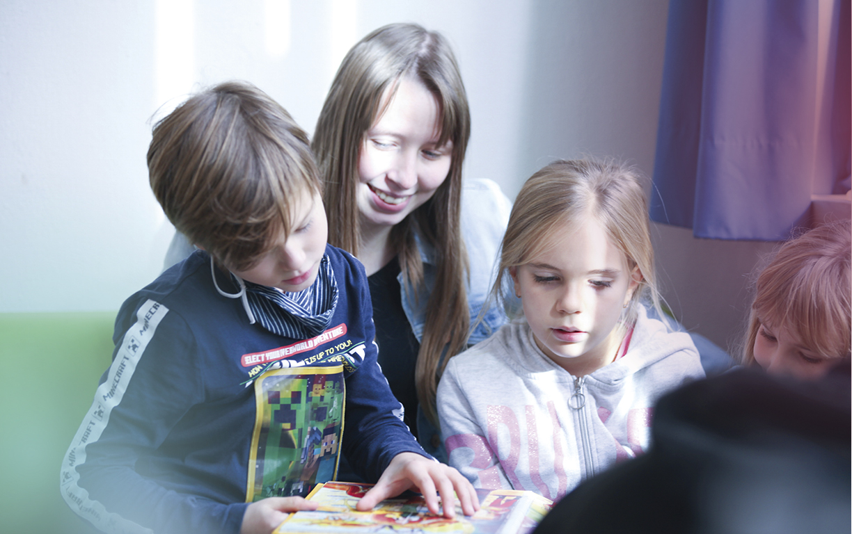 Eine junge Erzieherin sitzt auf einem Sofa und hat zwei Grundschulkinder auf ihrem Schoß, denen sie etwas vorließt.