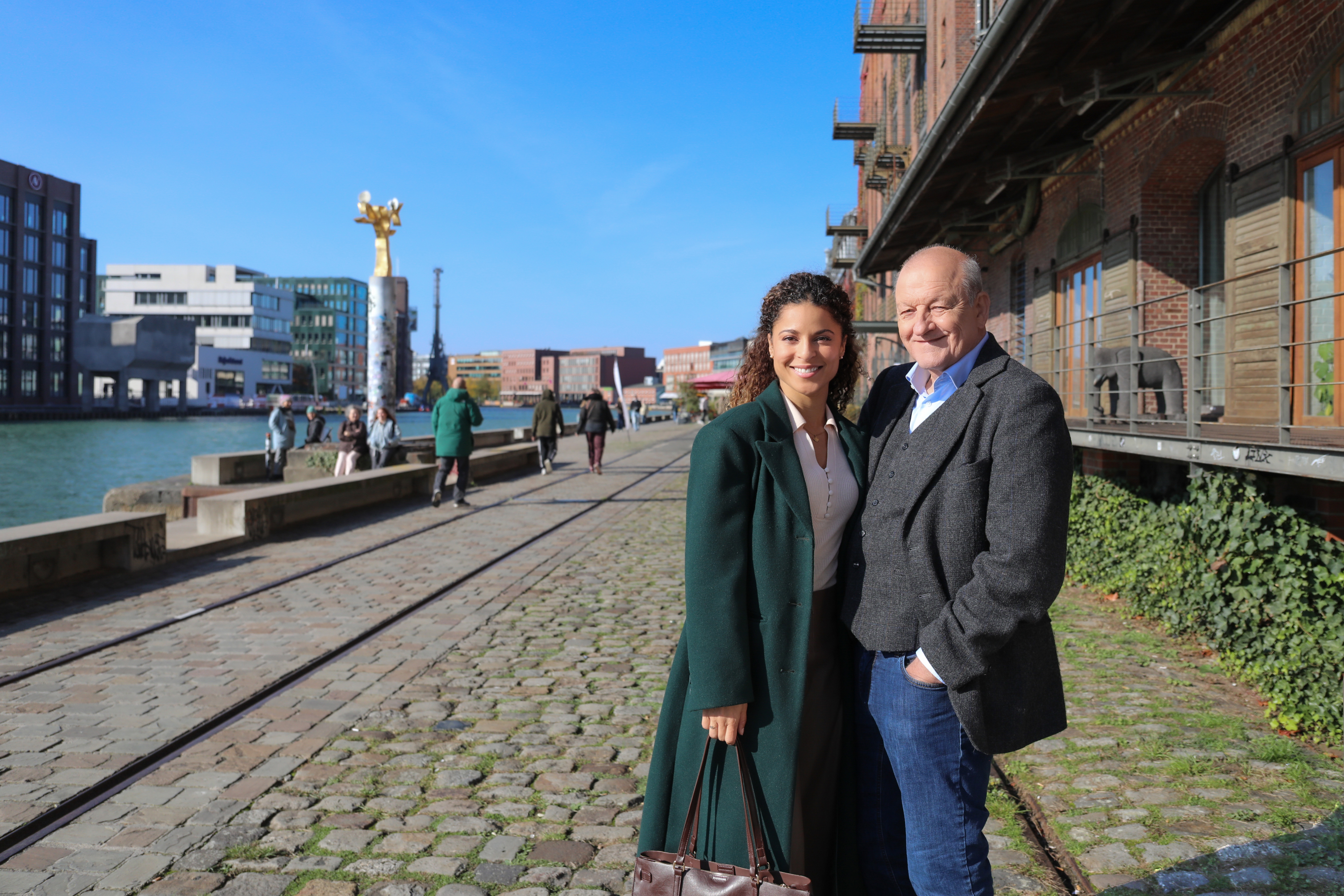 Die Darsteller Patricia Meeden und Leonard Lansink stehen nah beieinander vor einem Hafengebäude. Links ist das Hafenbecken zu sehen.