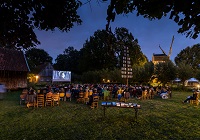 Blick auf das Gelände des Mühlenhofs in abendlicher Dämmerung, im Hintergrund ist die Film-Leinwand zu sehen, vor der viele Menschen auf Stühlen sitzen.