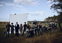 Eine Gruppe von Menschen, erkennbar als Filmteam steht mit Kamera und anderen filmtechnischen Geräten auf einer großen Waldlichtung