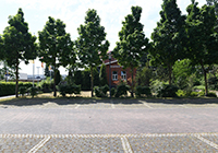 Der alte Bahnhof Reken - Parkplatz neben dem Bahnhofsgebäude mit Blick auf den rot geklinkerten Bahnhof durch dichte grüne Laubbäume