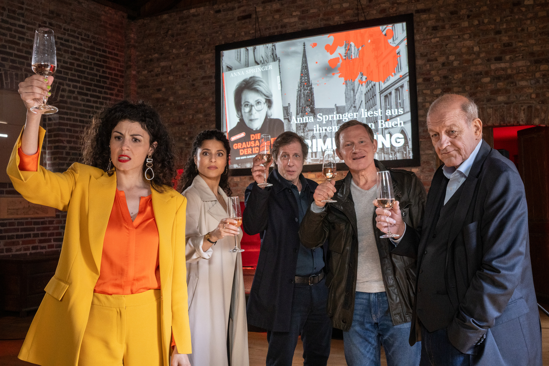 "Wilsberg – Blut geleckt": Lucia Hillbeck (Liza Tzschirner, v.l.), Dr. Tessa Tilker (Patricia Meeden), Ekki (Oliver Korittke), Overbeck (Roland Jankowsky) und Wilsberg (Leonard Lansink) stoßen auf das neue Buch von Hauptkommissarin Anna Springer (Rita Russek) an.