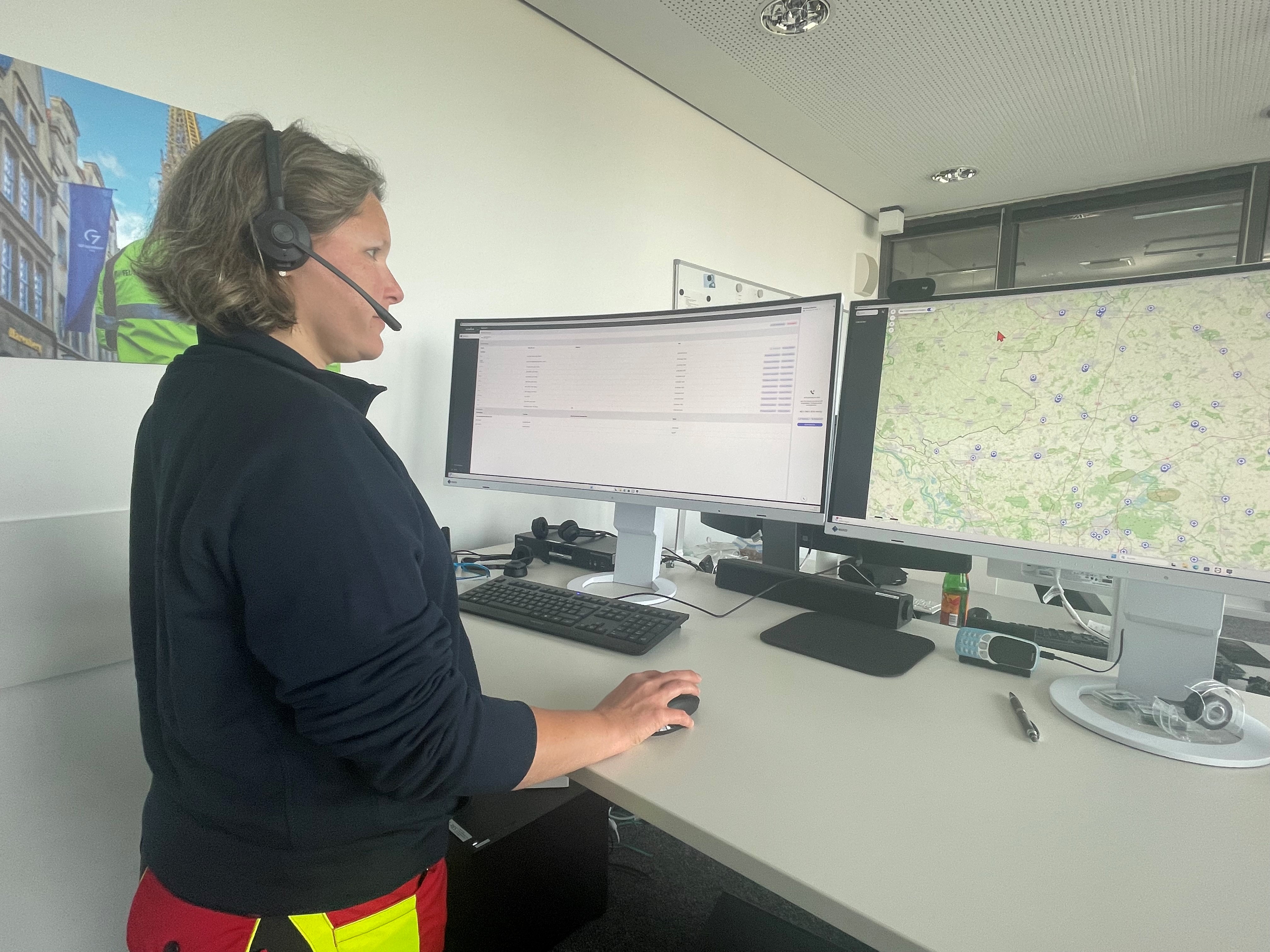 Eine Frau steht an einem Schreibtisch trägt ein Headset und guckt auf zwei Monitore.