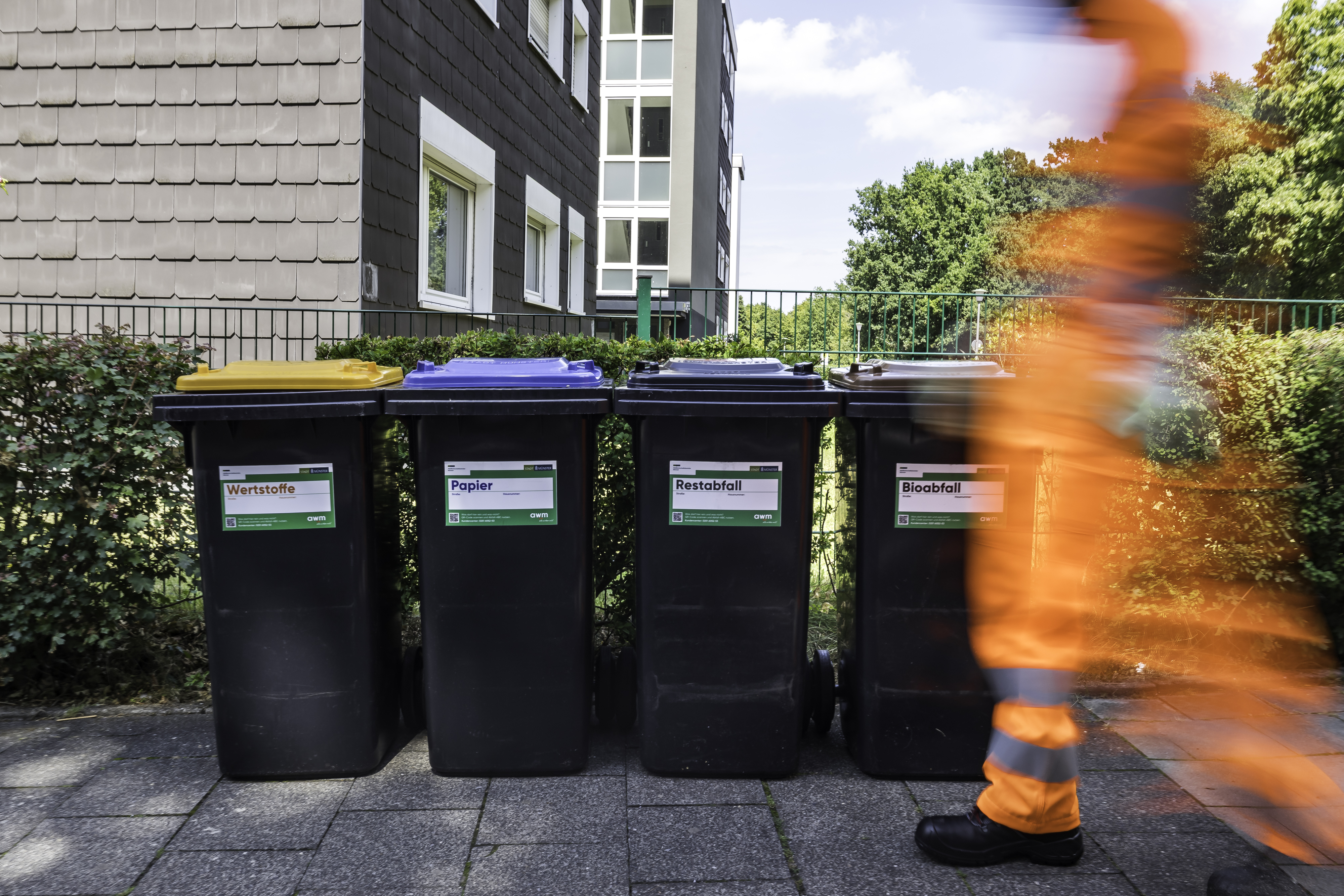 Vier Mülltonnen vor einem eingezäunten Grundstück, im Vordergrund ein schemenhafter Mitarbeiter der awm