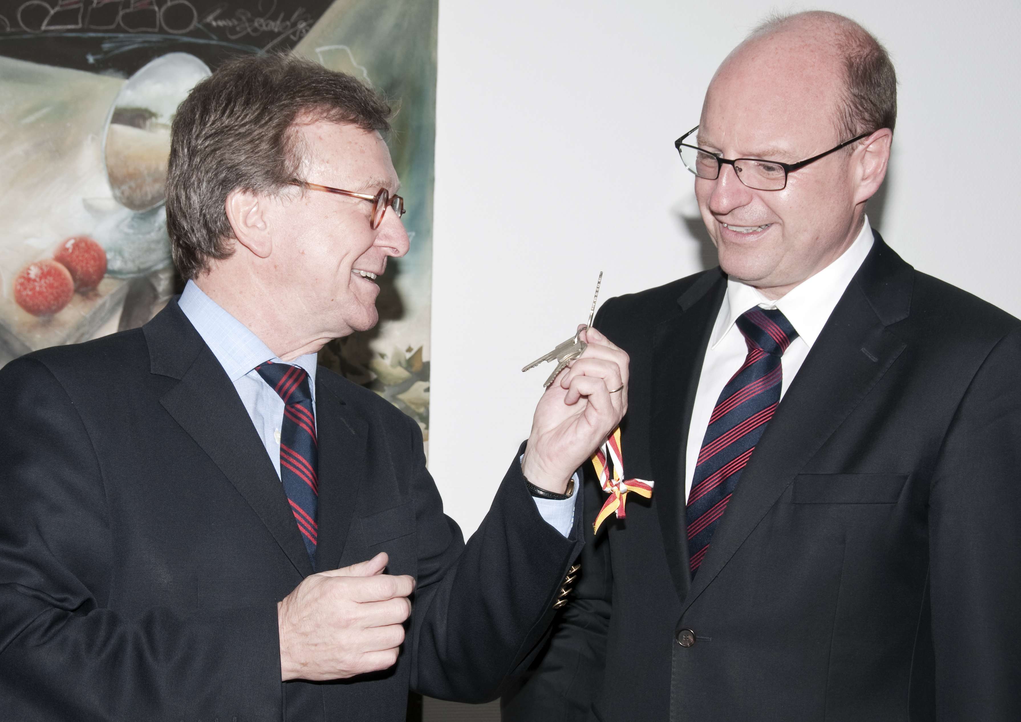 Ein Mann übergibt einem weiteren Mann einen Schlüssel. Beide Männer sind im Anzug und lächeln.