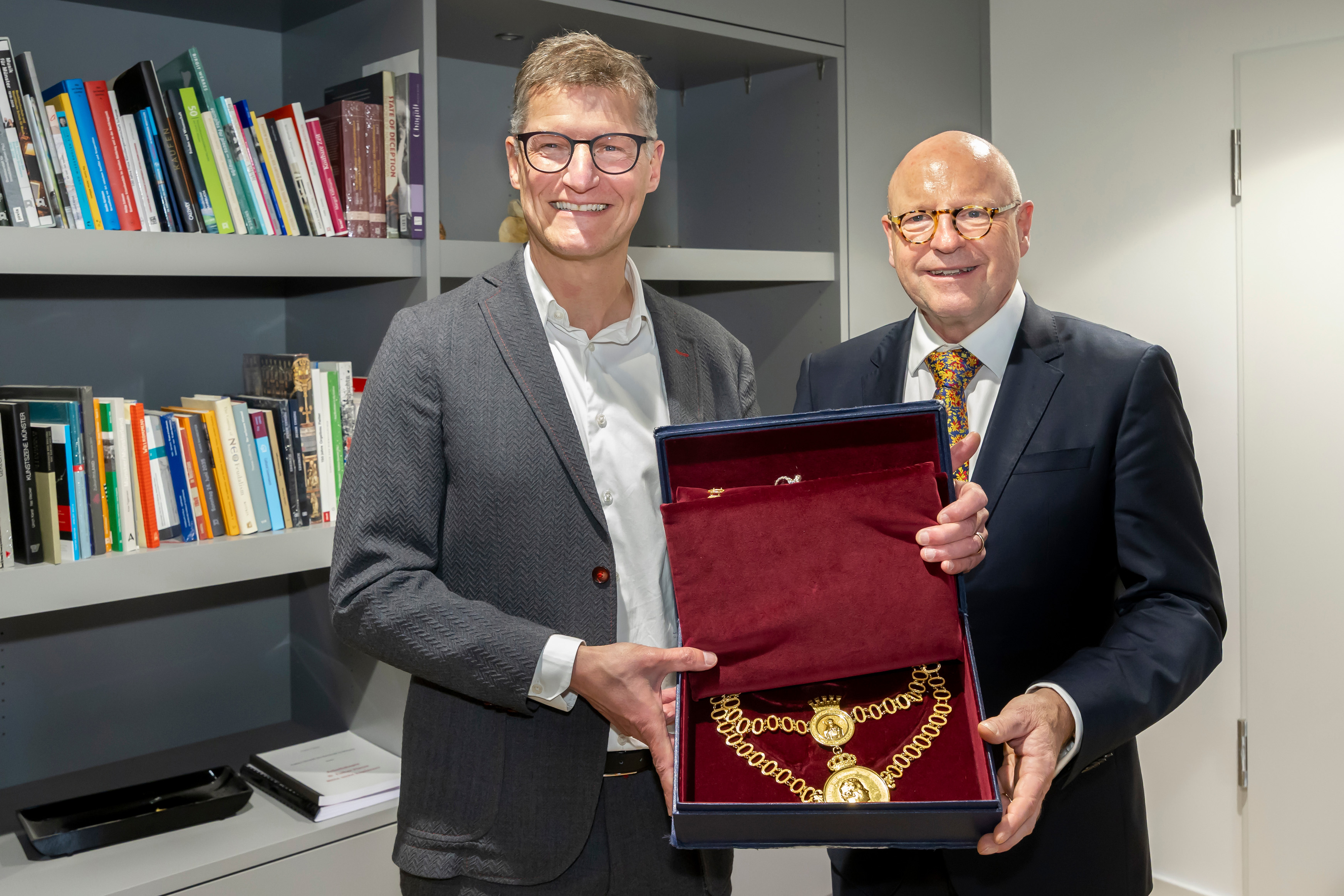 Zwei Männer lächeln in die Kamera und halten eine Schmuckschatulle mit einer großen Kette darin in den Händen