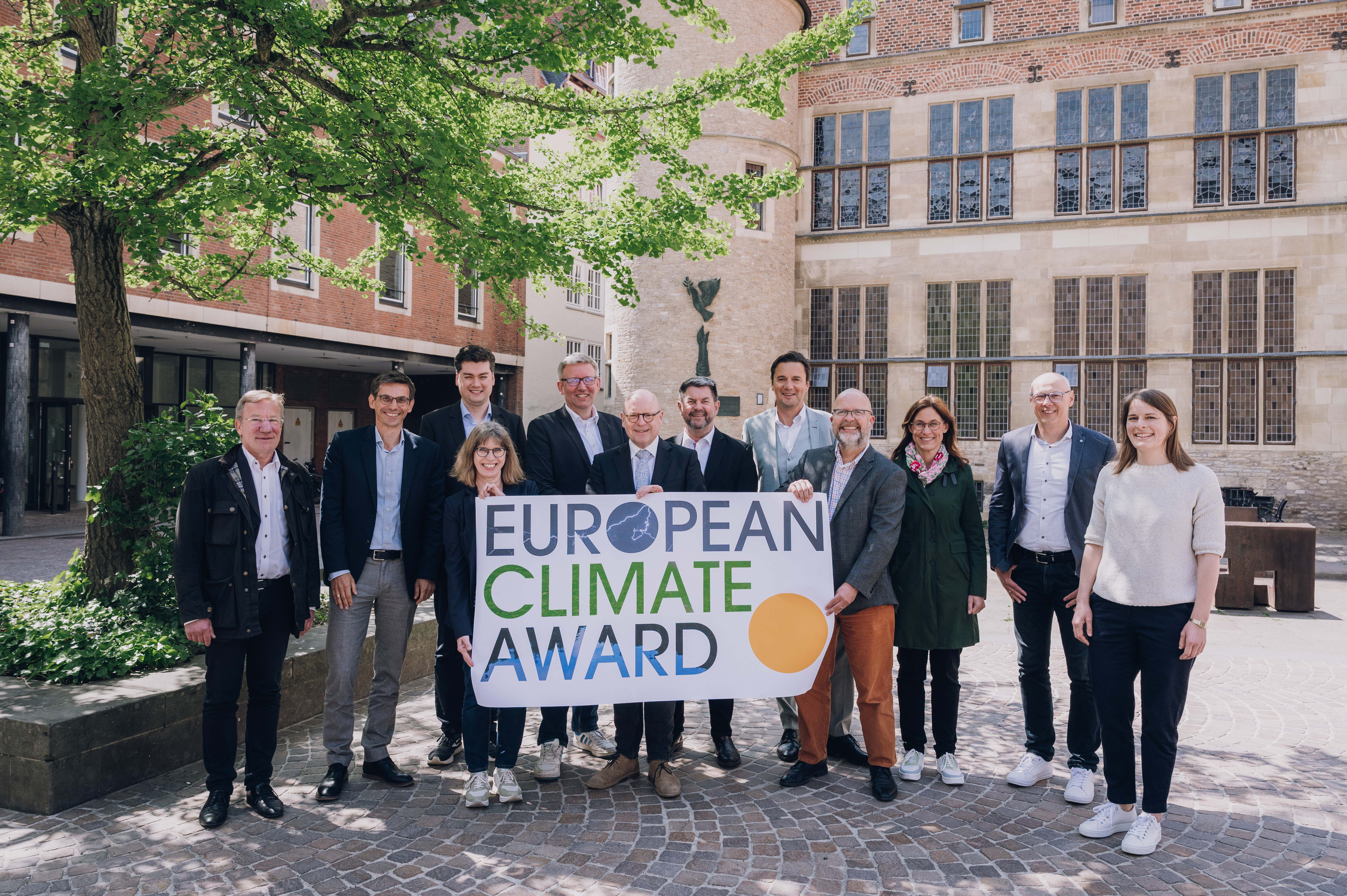 Die Menschengruppe steht im Schatten eines Baumes auf dem Rathausinnenhof und drei Personen in der Mitte halten ein mittelgroßes Banner mit der Aufschrift "European Climate Award" hoch.