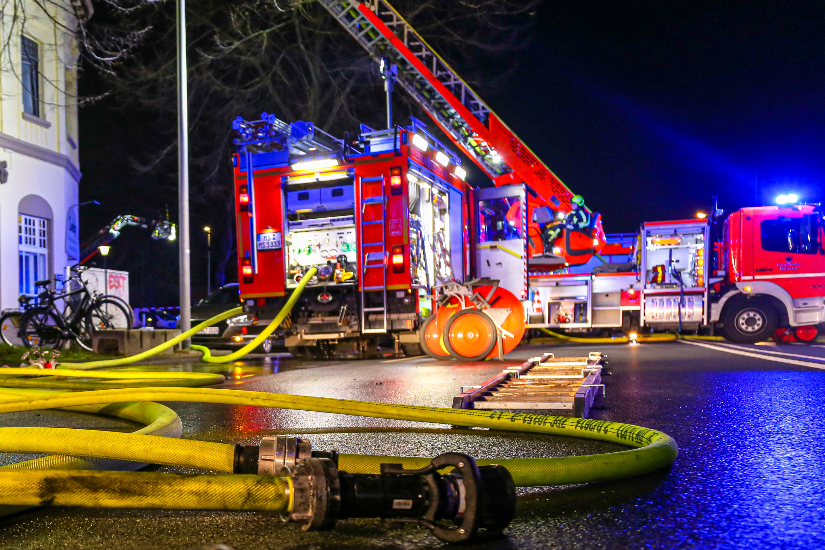Bei Nacht - Zwei Feuerwehrwagen mit Blaulicht im Hintergrund, vorne die Straße mit gelbem Feuerwehrschlauch