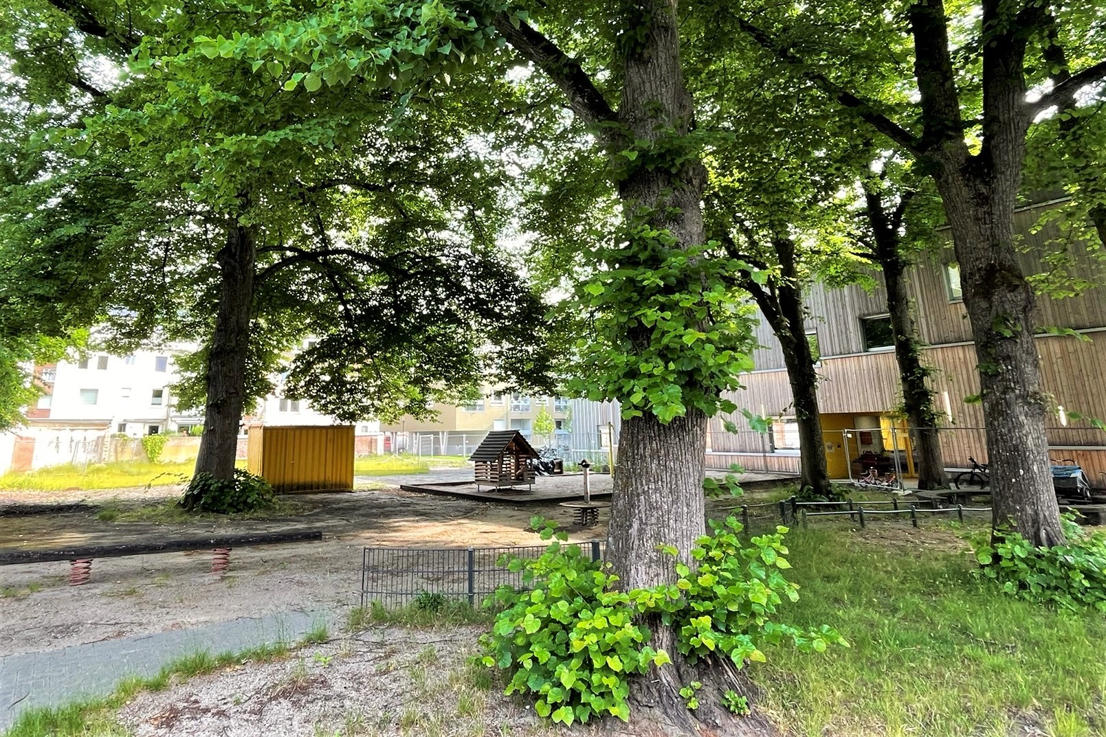Blick auf ein ehemaliges Schulgelände unter großen Bäumen, im Hintergrund das Schulgebäude. In der Bildmitte verlassene alte Spielgeräte und ein Sandkasten.
