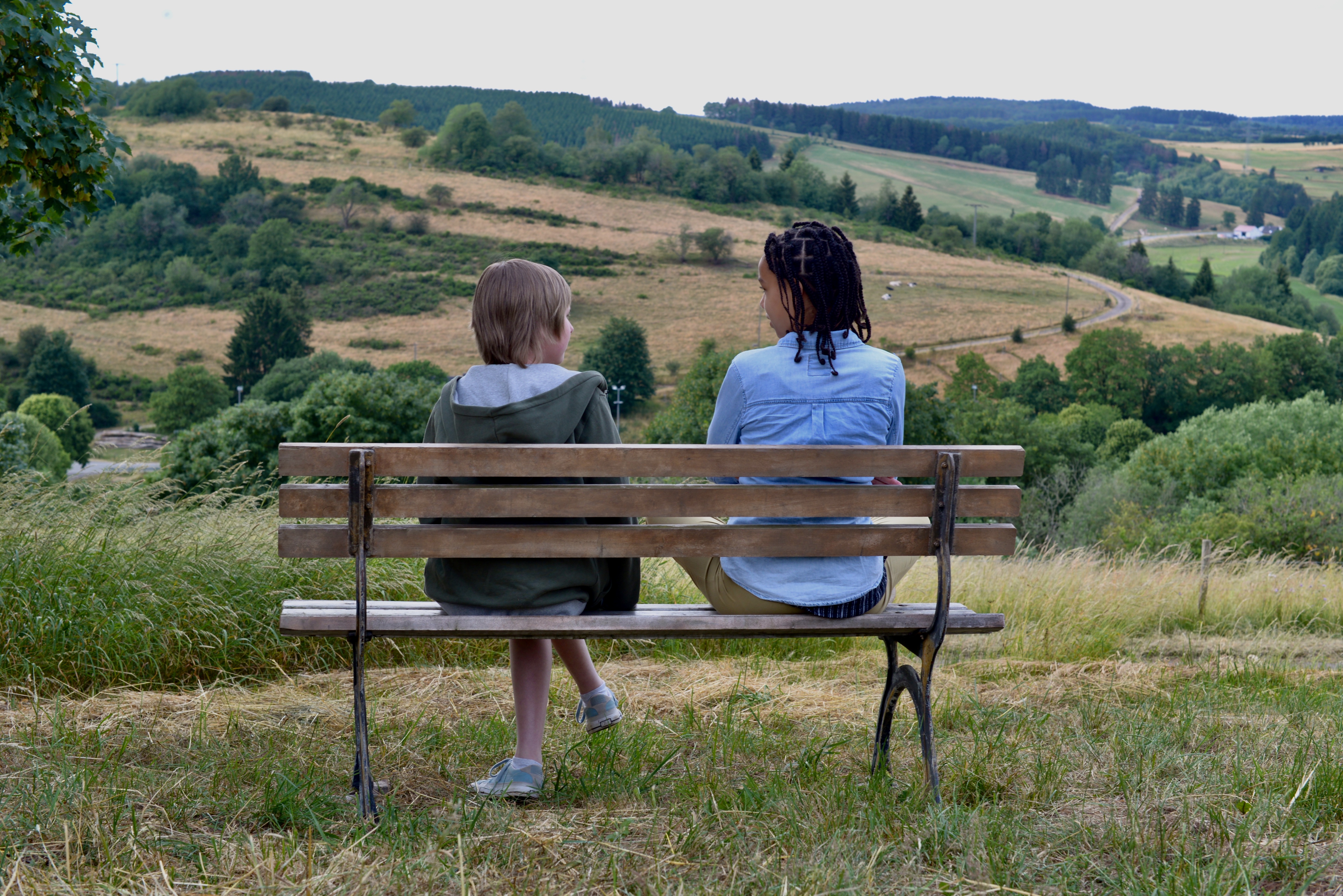 Zwei Kinder sitzen in einer Landschaft auf einer Bank, von hinten fotografiert.