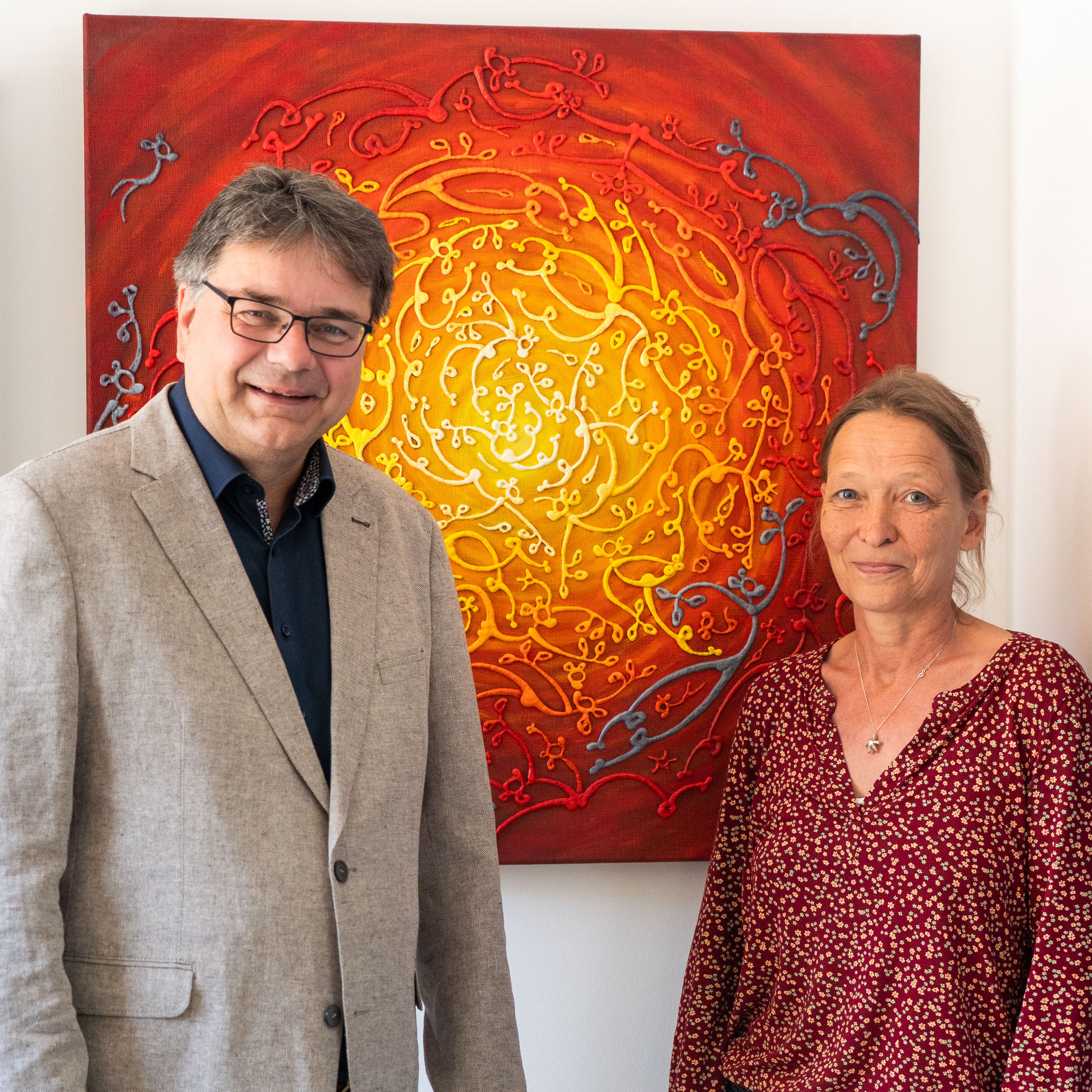 Ein Mann (links im Bild) und eine Frau stehen vor einem Gemälde in leuchtend gelb-roten Farben mit kreisförmigen Ornamenten