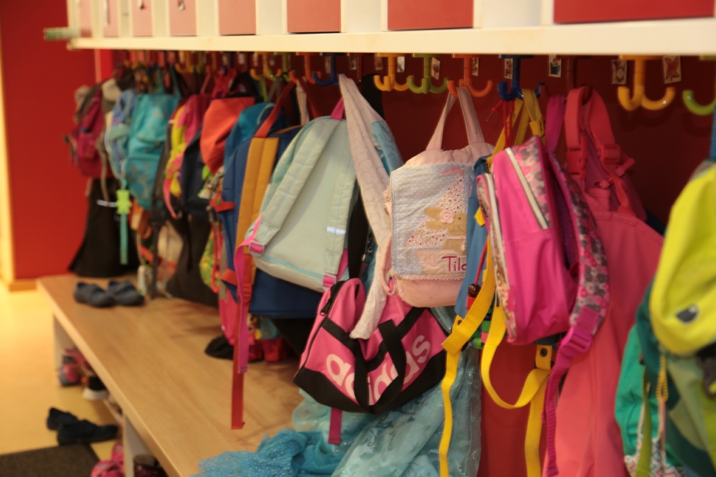An einer Garderobe in einer Kita hängen Kinderrucksäcke und Kinderjacken in unterschiedlichen Farben.