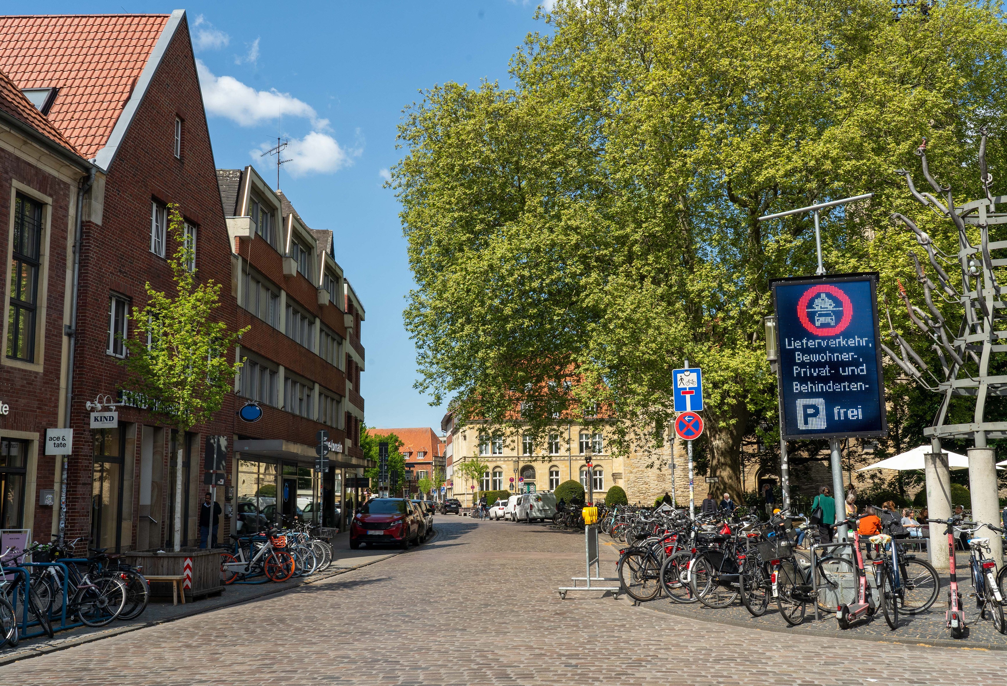 Sicht auf bzw. in die geplasterte Königgstraße bei sonnigem Wetter. Am Straßenrand viele Fahrräder, rechts am Straßenrad steht eine große LED-Tafel, deren Anzeige die Zufahrt beschränkt.