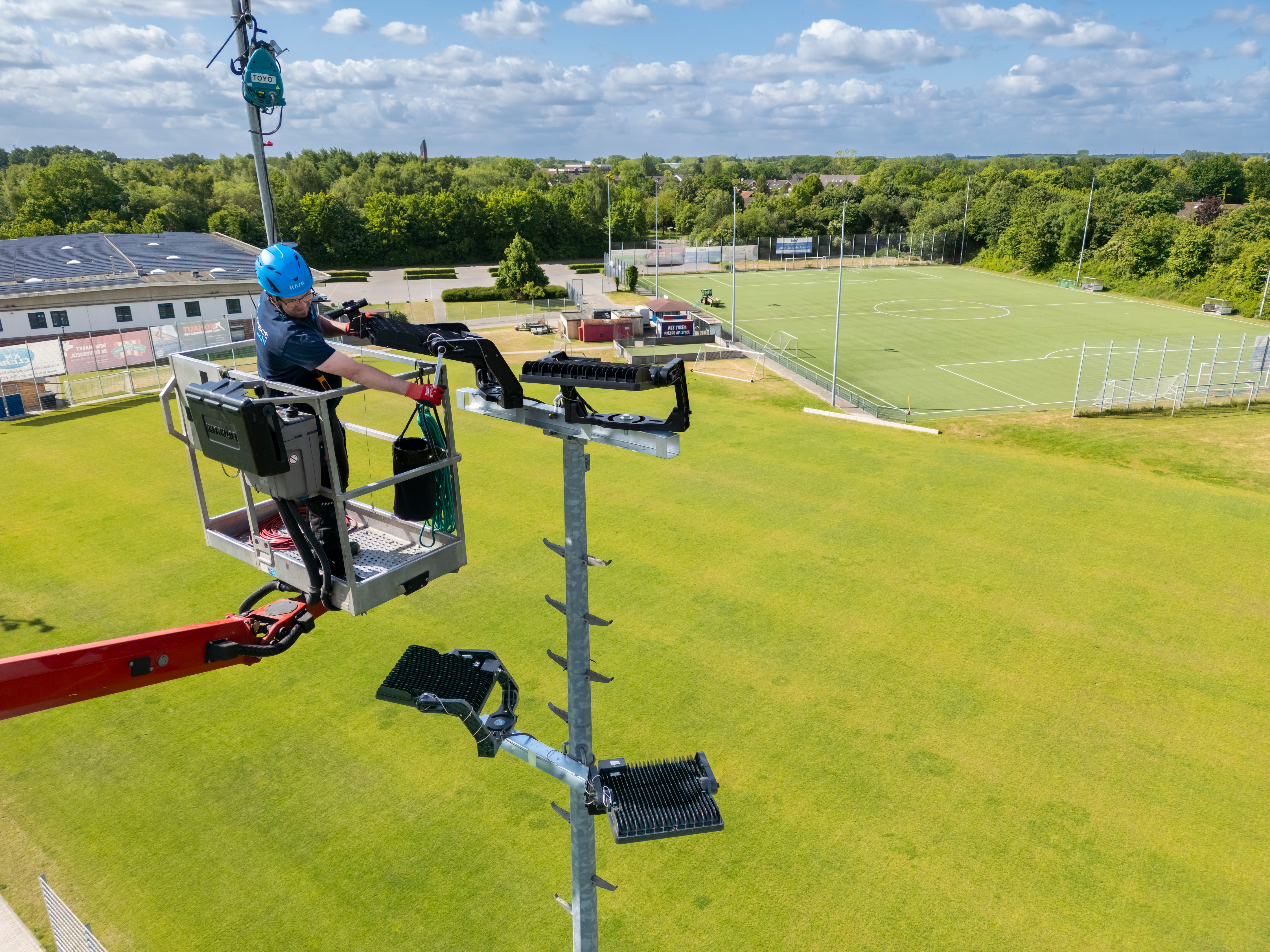 Ein Techniker im Korb eines Hubwagens arbeitet an einem Flutlicht, unter ihm der Sportplatz, im Hintergund sind Wald und der Horizont zu sehen.