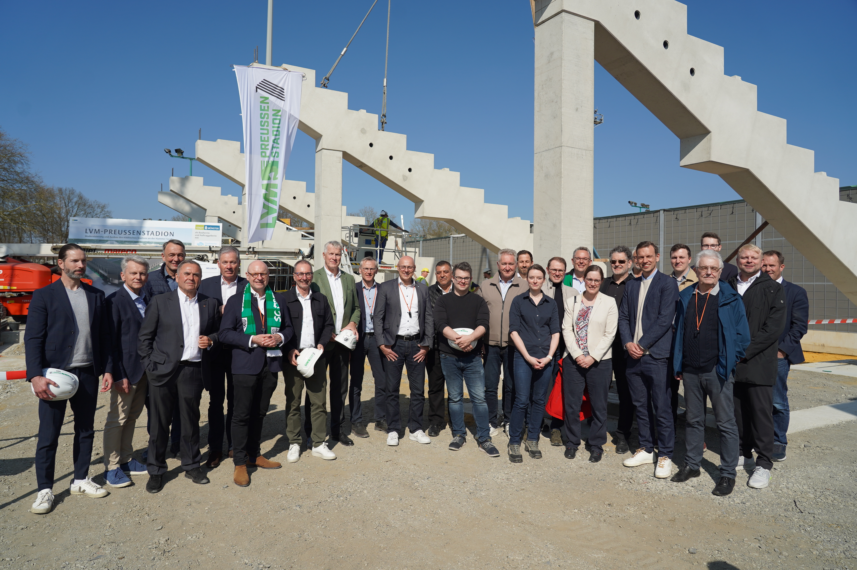 Eine große Gruppe Personen steht aufgereiht vor den ersten platzierten Zahnbalken der Westtribüne und schaut in die Kamera.