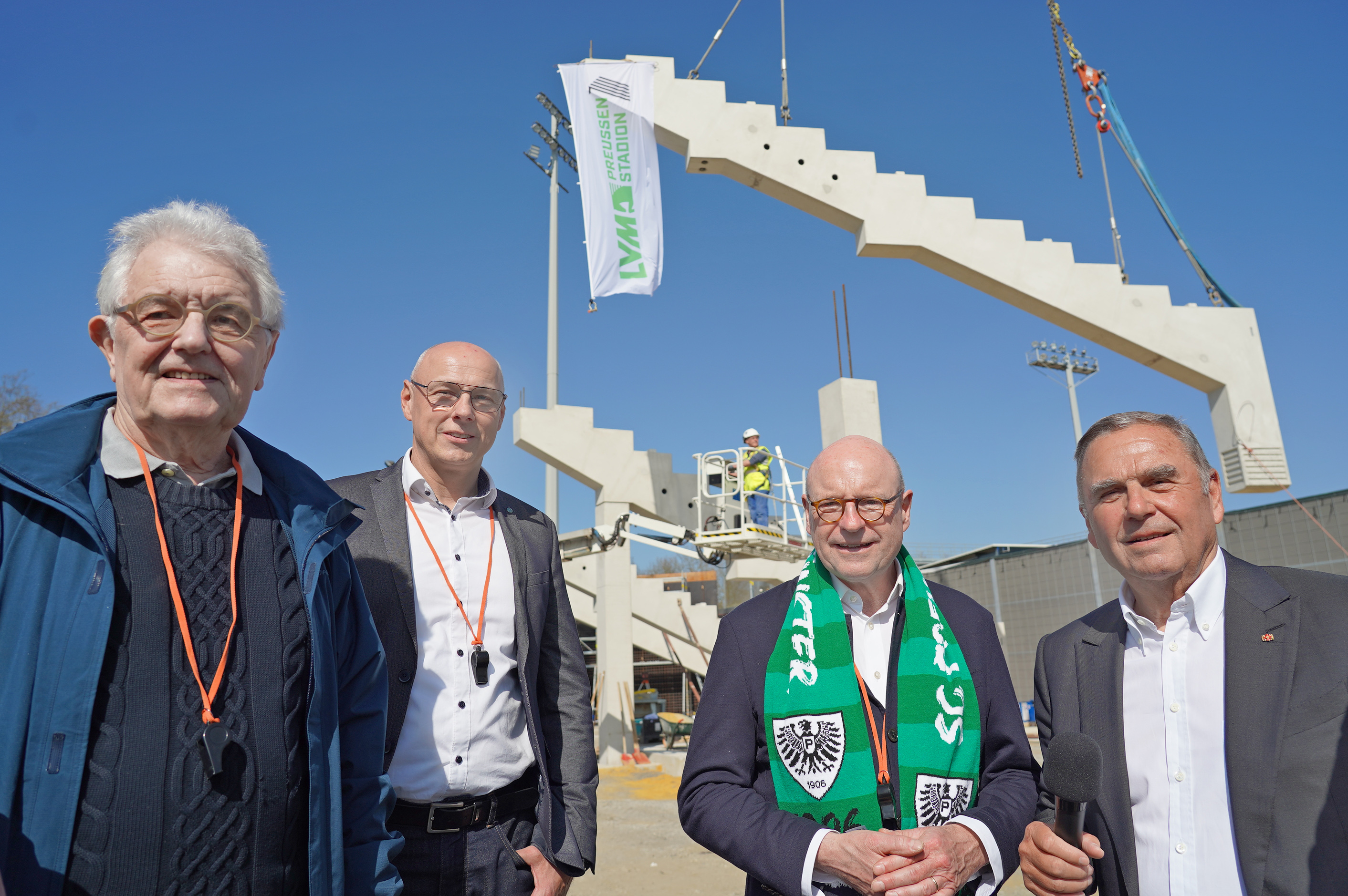 Christoph Strässer (v.l.), Frank Gäfgen, Markus Lewe und Walter Hellmich stehen auf der Baustelle, hinter ihnen die ersten platzierten Zahnbalken der Tribüne und in Luft schwebt noch ein Zahnbalken am Kran.