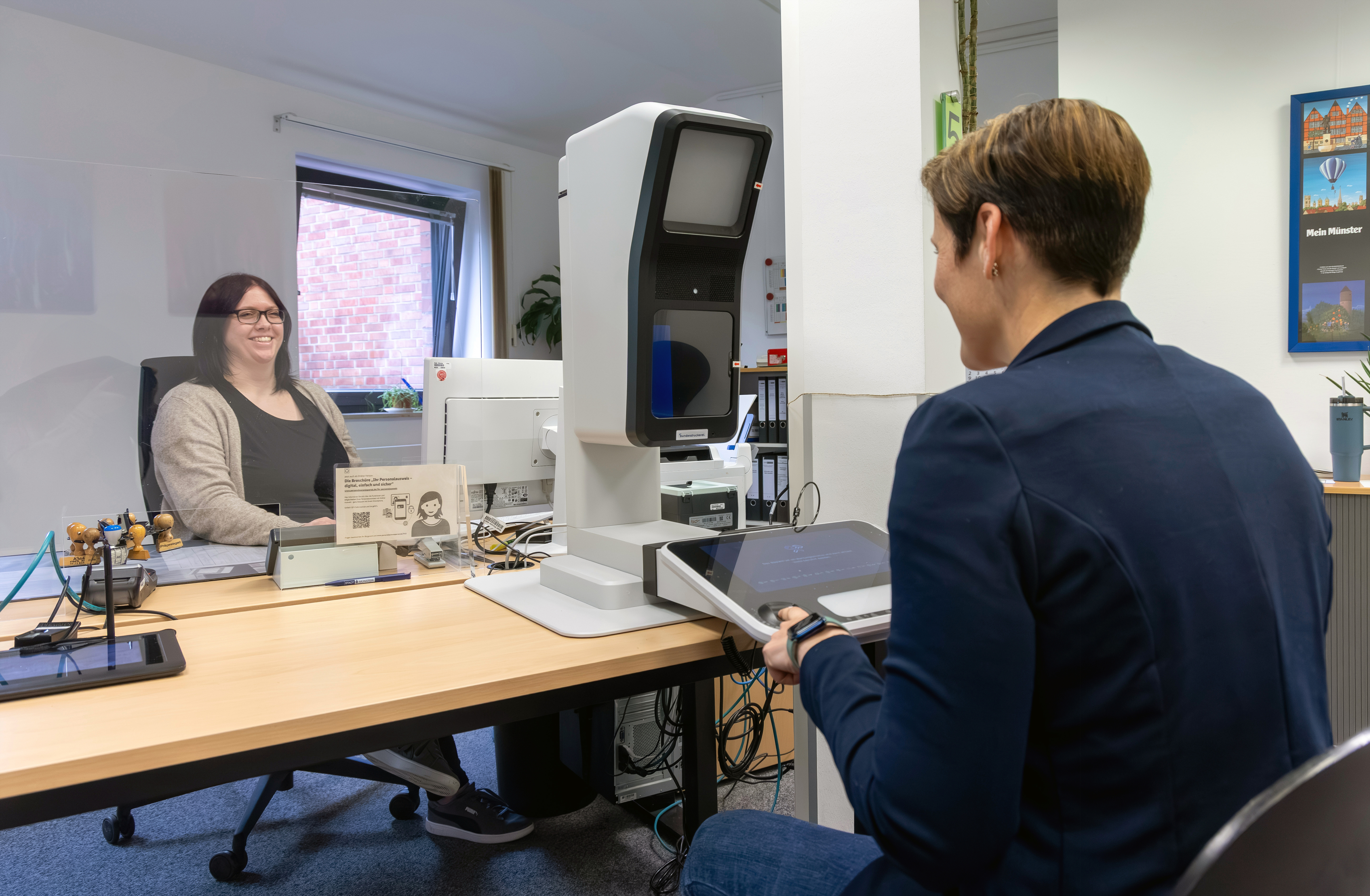 Ein Mann sitzt am Tisch vor dem Passbildautomaten. Hinter dem Tisch sitzt eine Mitarbeiterin des Bürgerbüros.