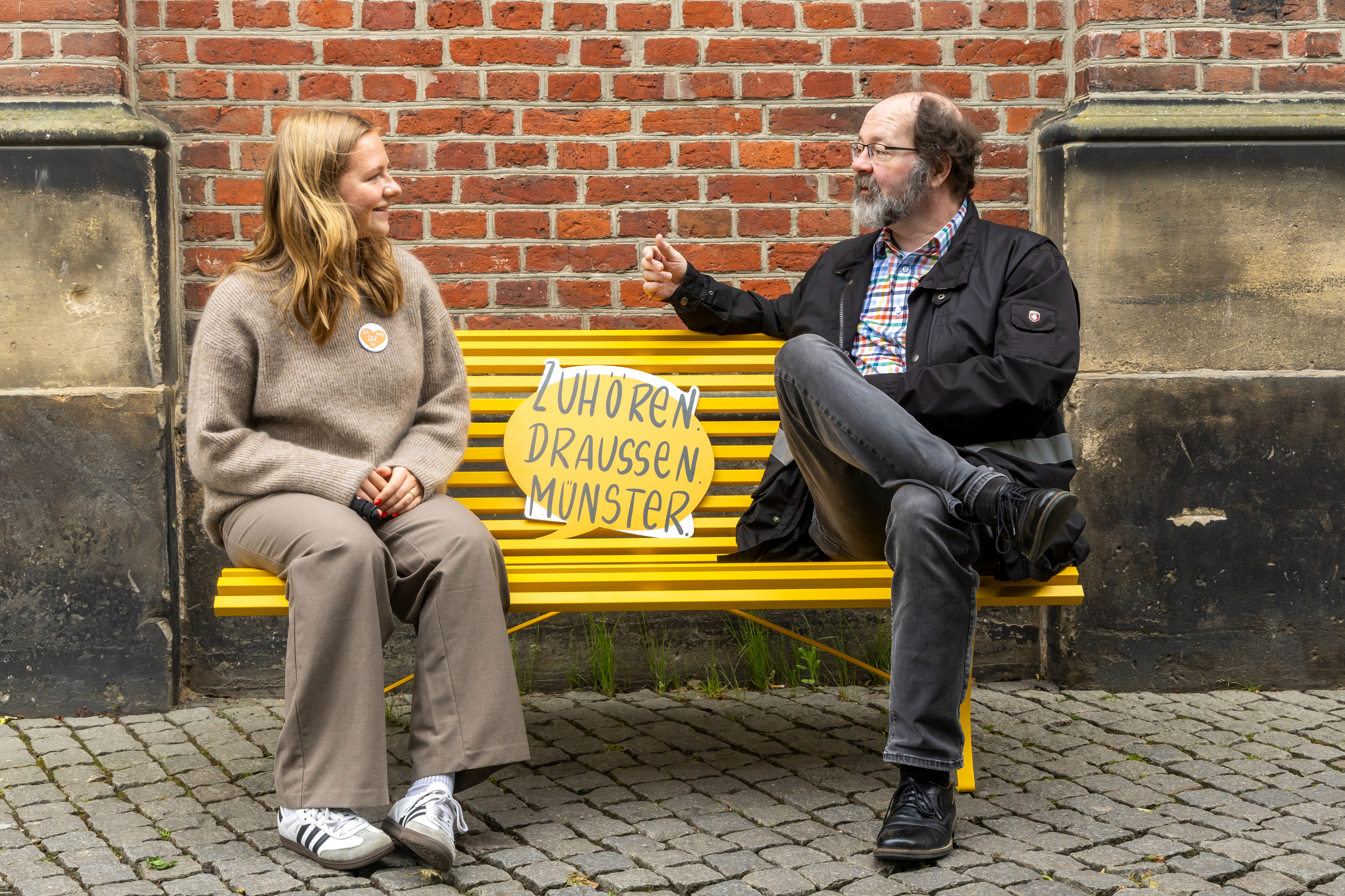Die zwei ehrenamtlichen Zuhörer Sara Stetter und Dr. Achim Detering sitzen auf der gelben Bank