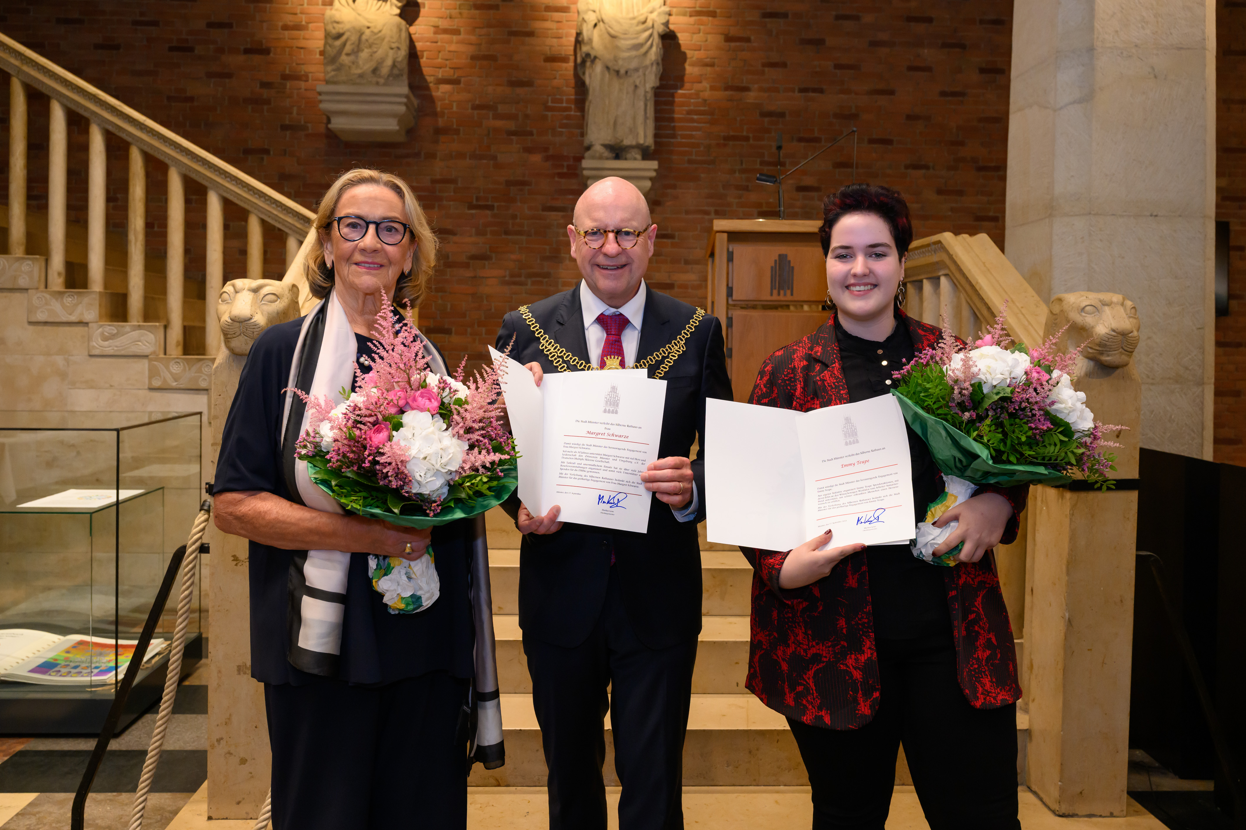 Die drei Personen stehen in der Bürgerhalle des Rathauses und halten Urkunden und Blumensträuße in den Händen.