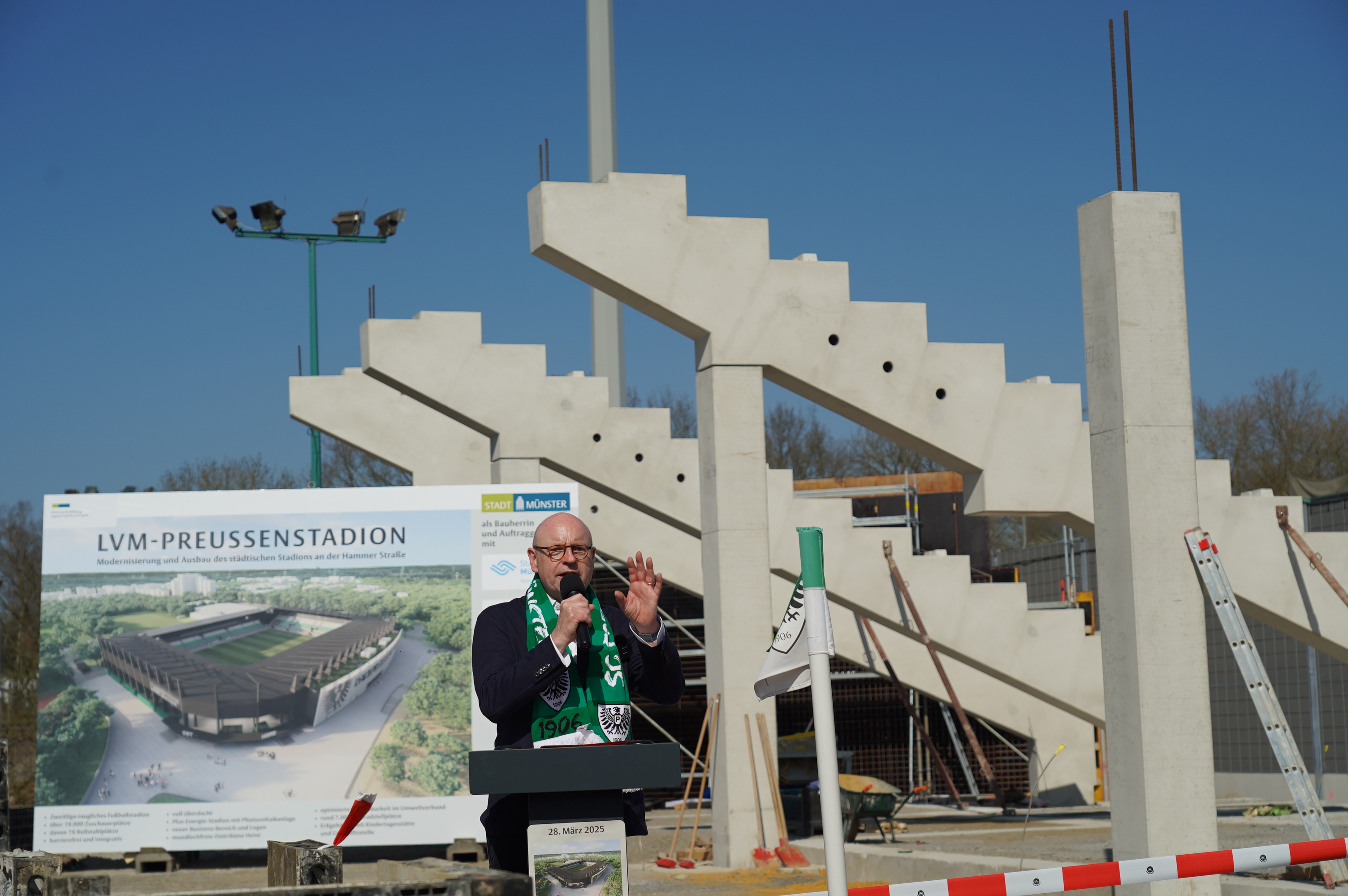 Ein Mann kniet auf einer Bühne. Im Hintergrund die ersten Stehlen eines Stadionneubaus