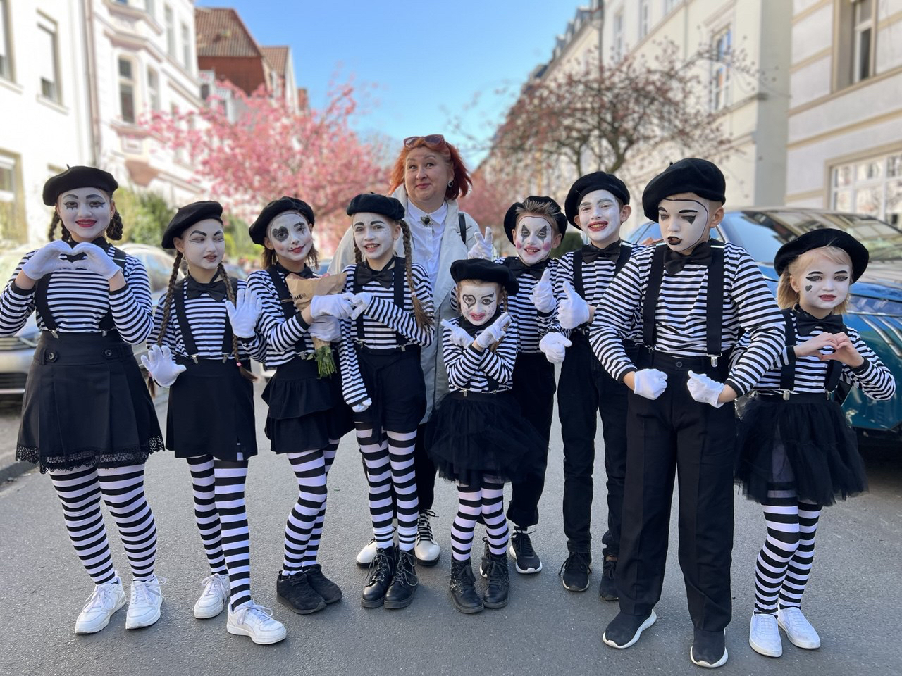 Neun Kinder als Pantomimen verkleidet und geschminkt stehen aufgereiht auf einer Straße, dahinter eine erwachsene Frau ohne Verkleidung oder Schminke. Im Hintergrund reihen sich links und rechts geparkte Autos, Häuserreihen und blühende Bäume.