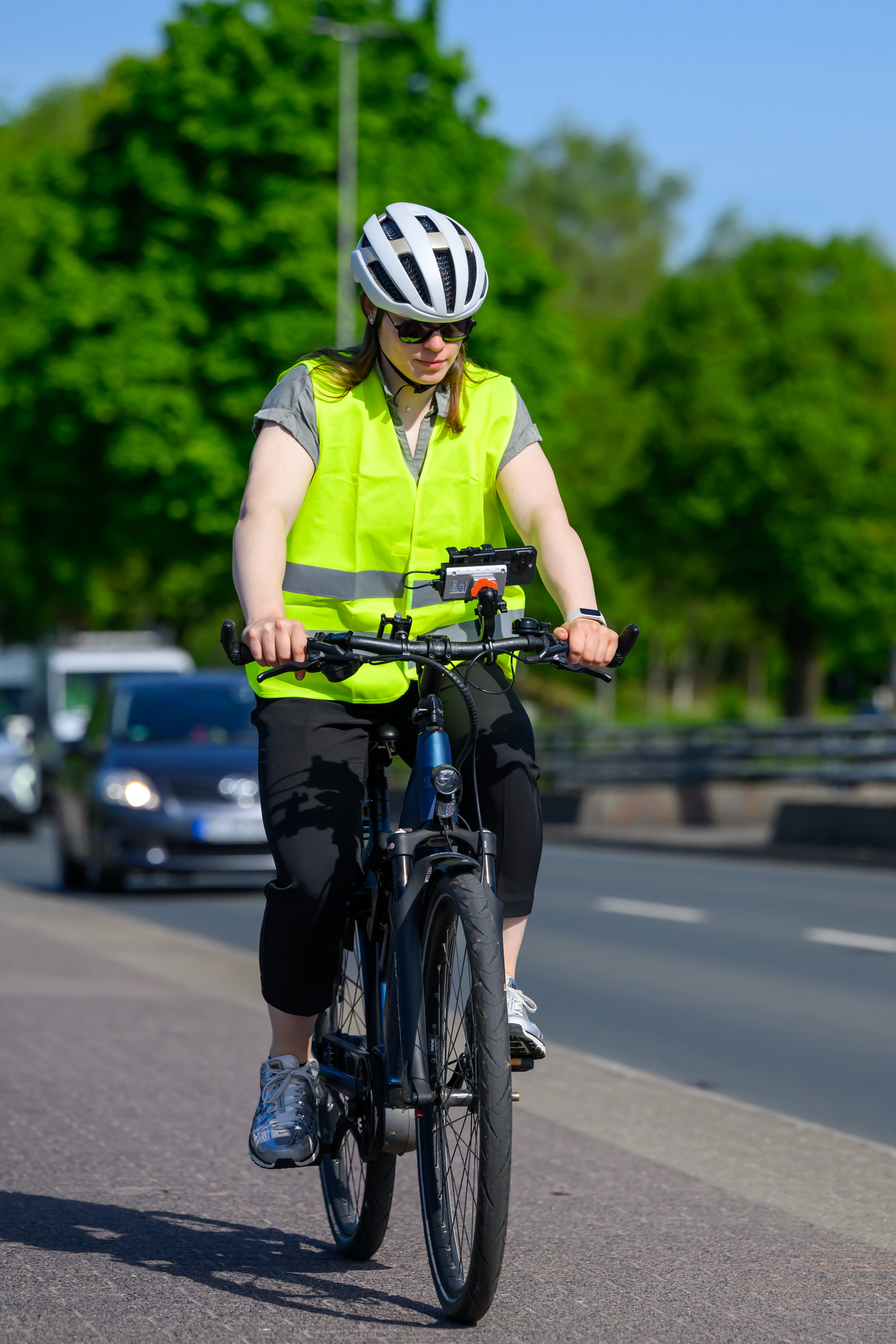 Eine Frau mit Sonnenbrille, Helm und Warnweste fährt auf einem Fahrrad einen Radweg entlang. Am Lenker ist ein Smartphone montiert, auf das die Radfahrerin zu schauen scheint. Im Hintegrund die Straße mit Autos, dahinter Bäume.