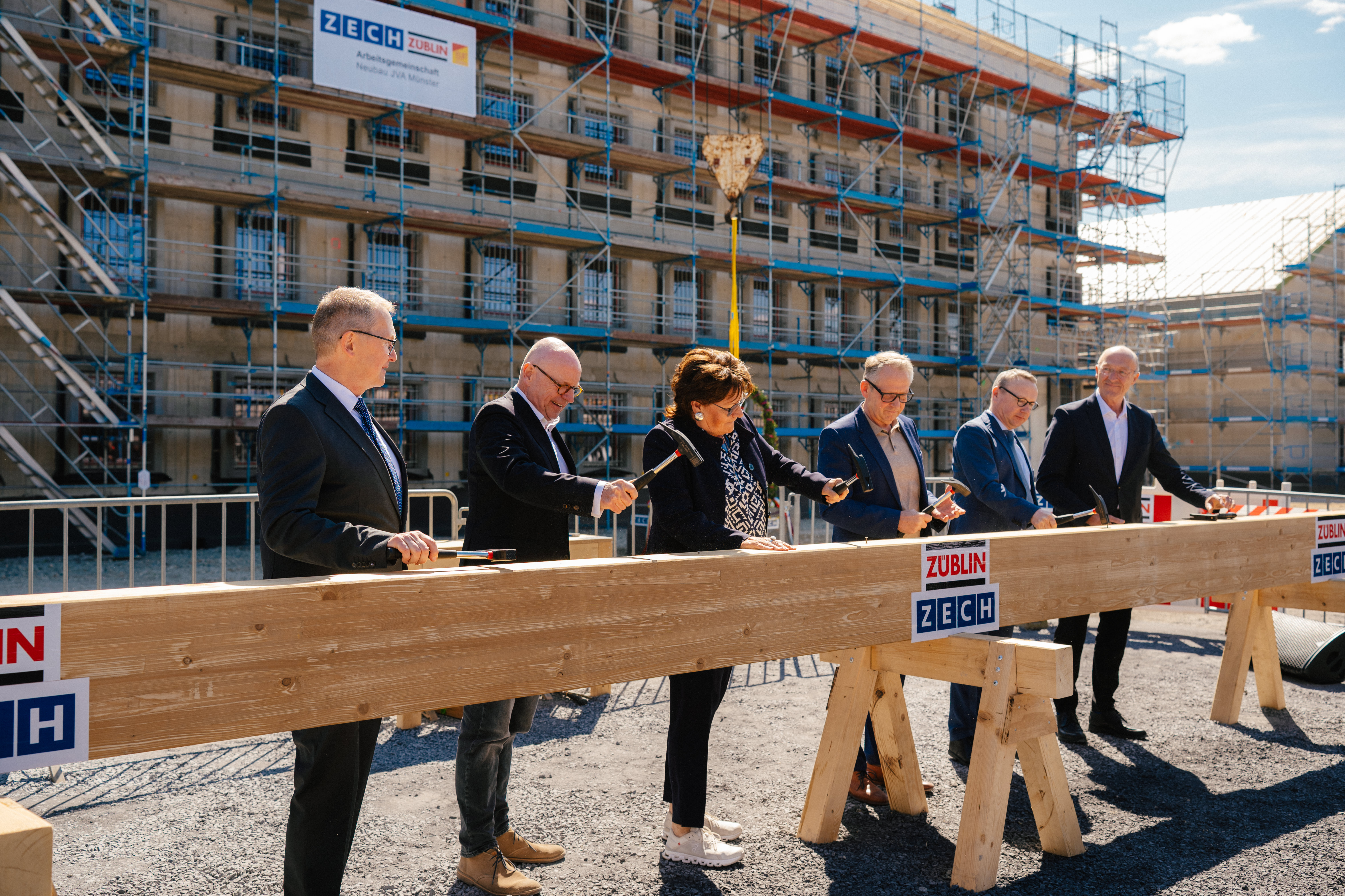 Die sechs Personen stehen vor dem mit dem Baugerüsten umhüllten Roh- bzw. Neubau der JVA und schlagen mit Hämmern jeweils einen Nagel in einem großen Dachbalken, der auf Holzböcken vor ihnen liegt.