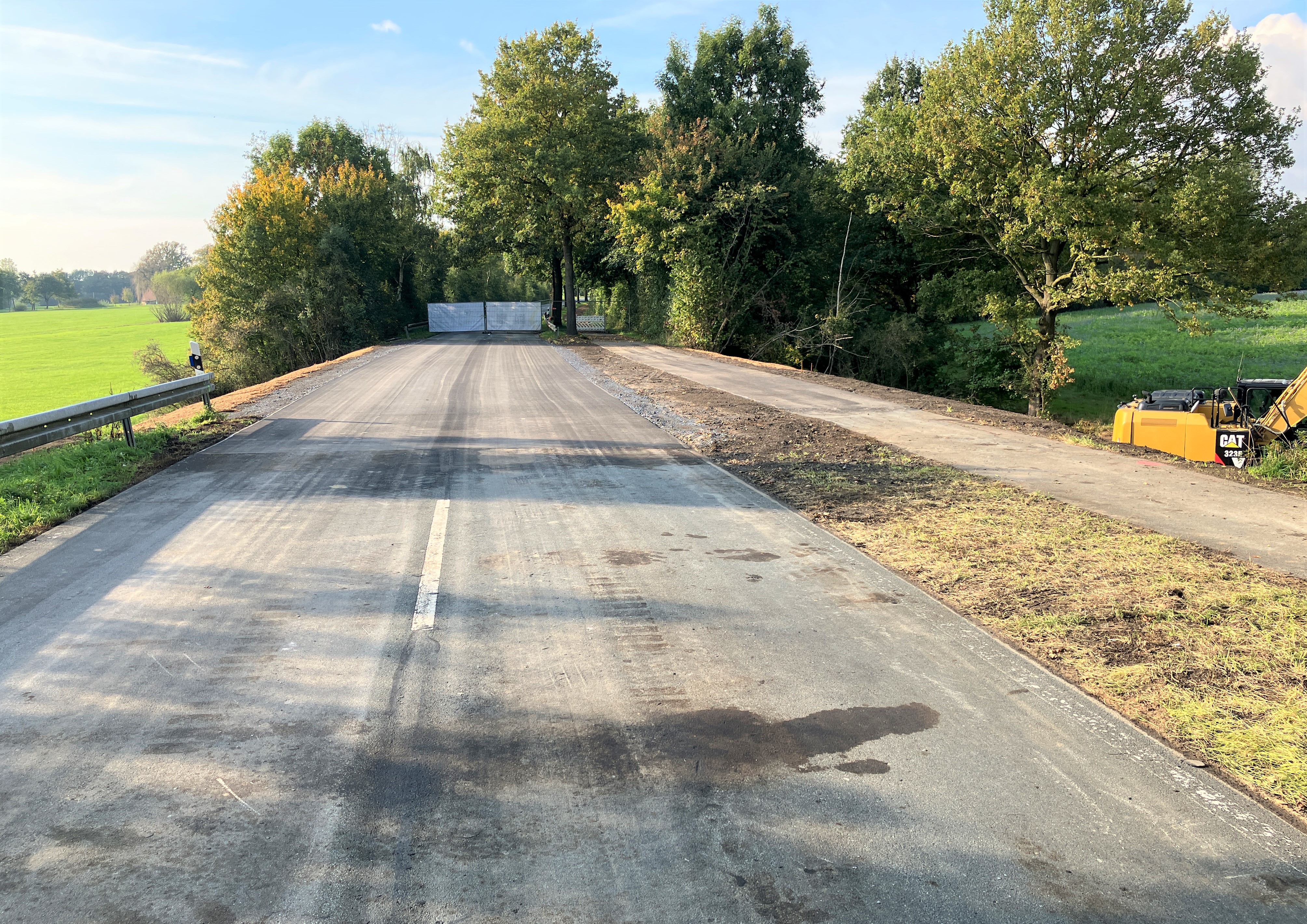 Leere Straße führt durch Wiesen und Bäume, am Rand steht ein Baufahrzeug