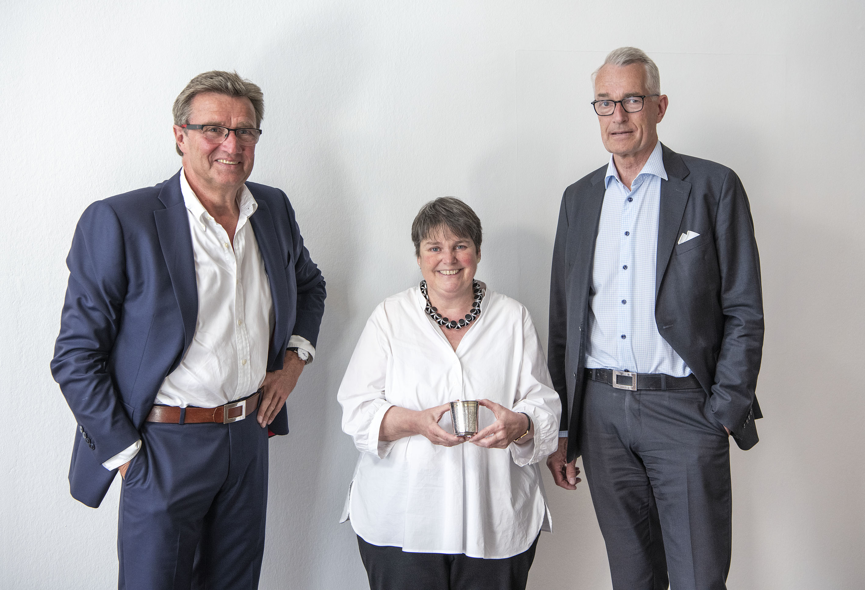 Links und rechts die beiden Herren, in der Mitte die Direktorin mit dem kleinen Silberbecher in der Hand. Alle vor weißer Wand.