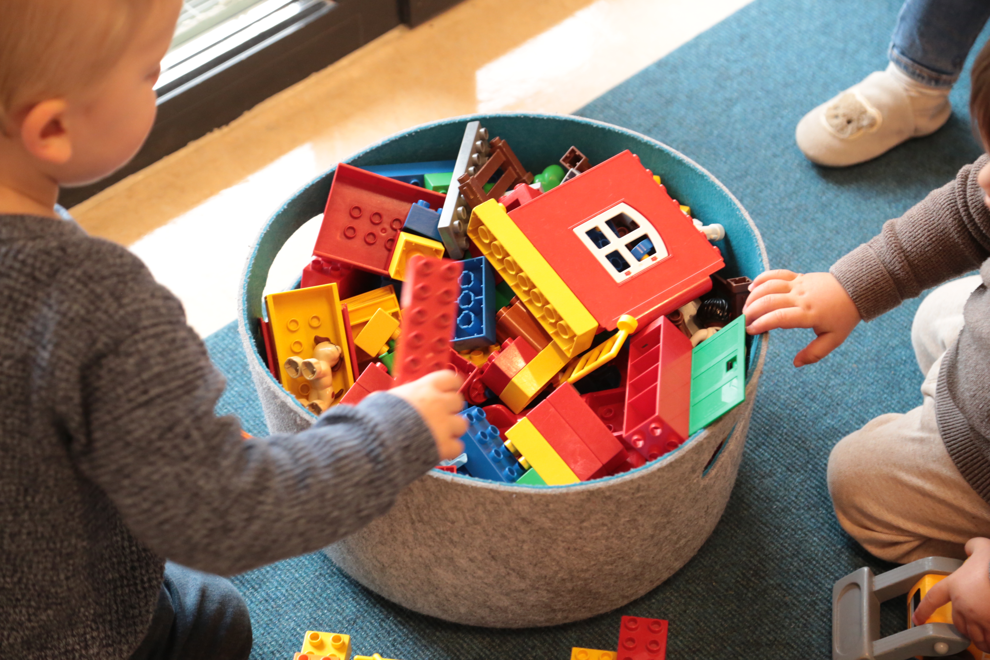 Im Fokus des Bildes ist eine grauer Filzkorb gefüllt mit Duplo-Steinen. Am Rand des Bildes sind Kinder zu erkennen. Eines von ihnen hat einen roten Duplo-Stein in der Hand.