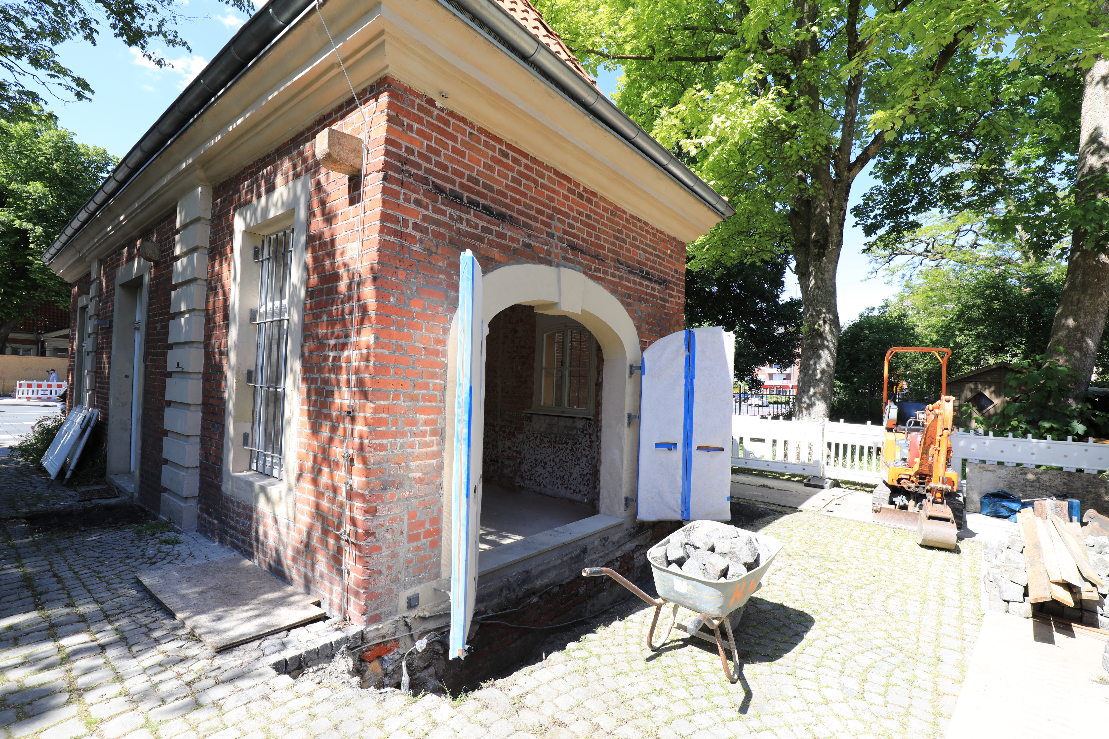 Ansicht des rot geziegelten Torhauses, mit geöffnetem Tor, davor eine Schubkarre mit Pflastersteinen. Im Hintergrund ein kleiner gelber Bagger.