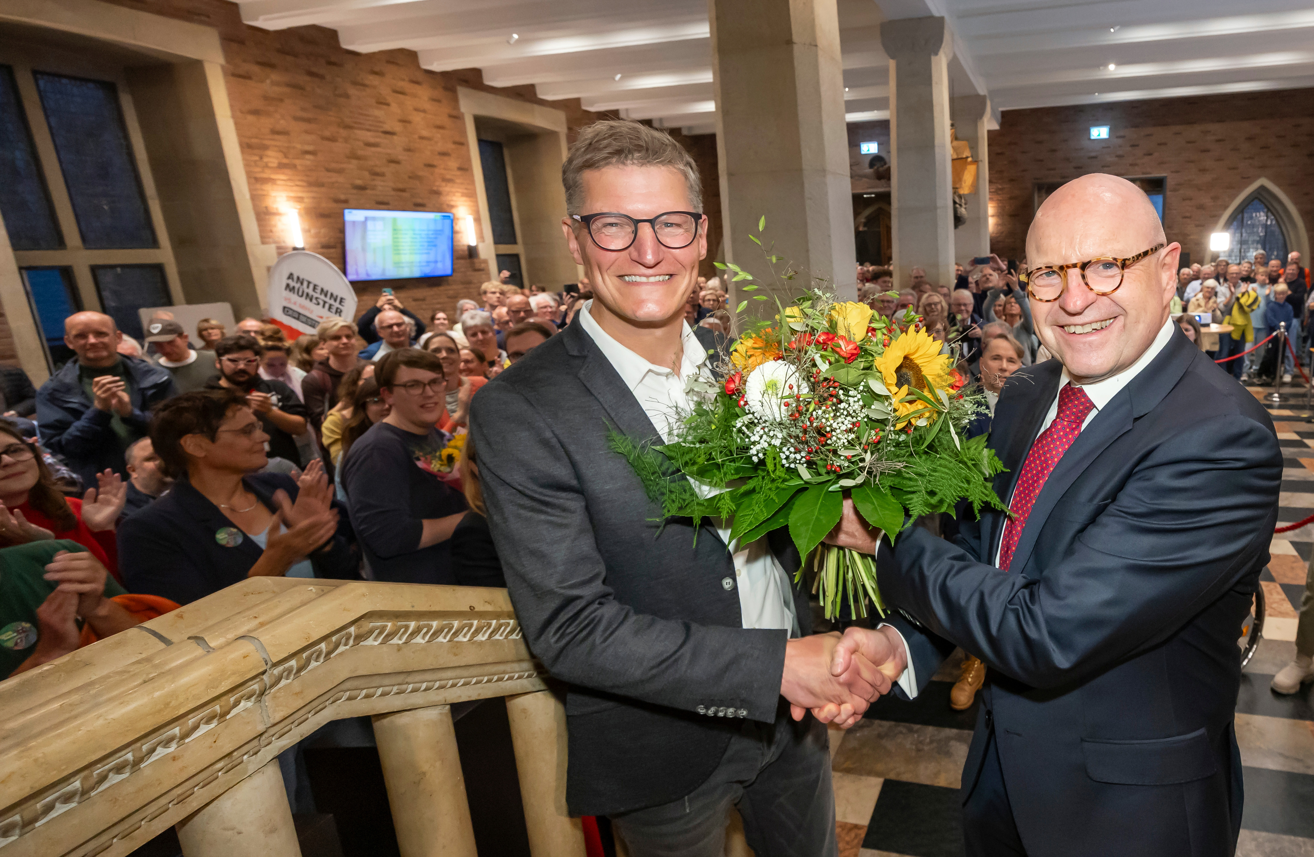 Tilman Fuchs und Markus Lewe mit Blumenstrauß in der gut besuchten Bürgerhalle im Historischen Rathaus in Münster
