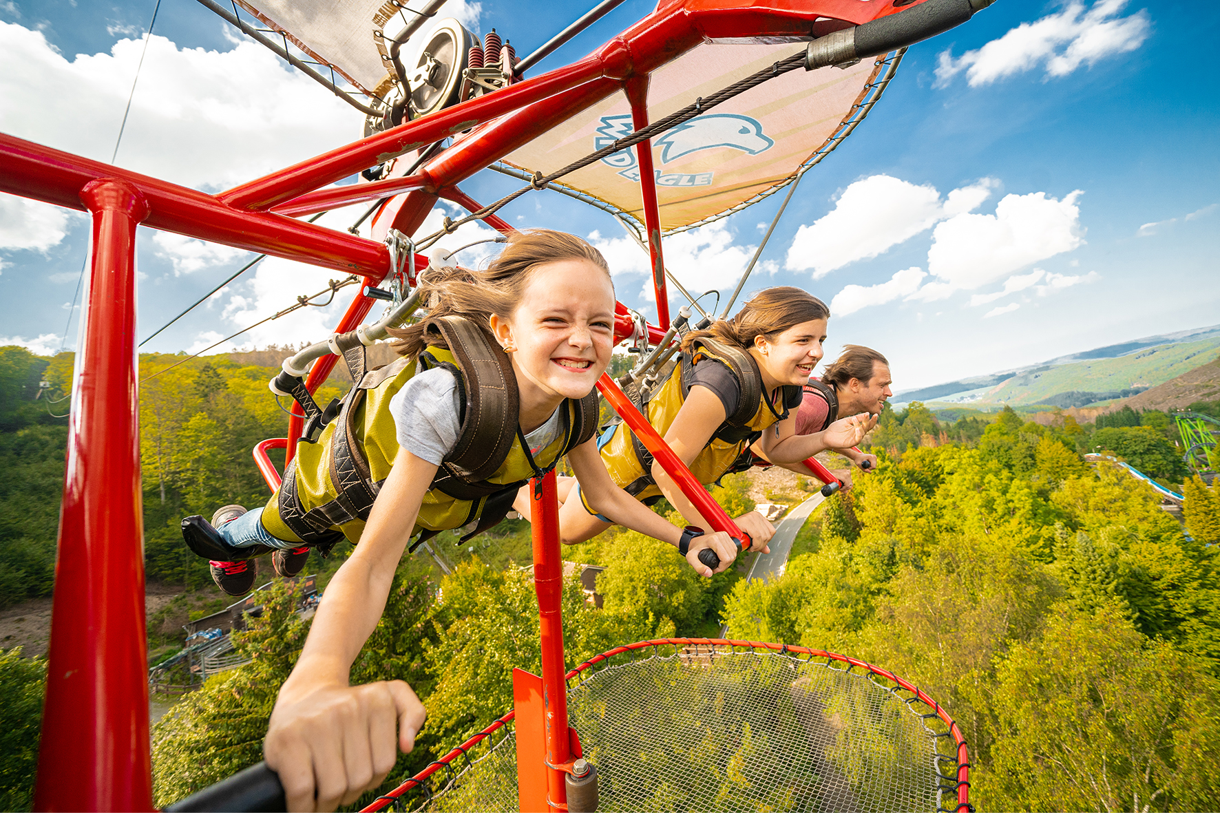 Zwei Mädchen und ein Erwachsener hängen in dem Fahrgeschäft/Drachenflieger "Wild Egale" im Freizeitpark Fort Fun. Über ihnen ist der Himmel, unter ihnen ein Sicherheitsnetz und ein Wald.