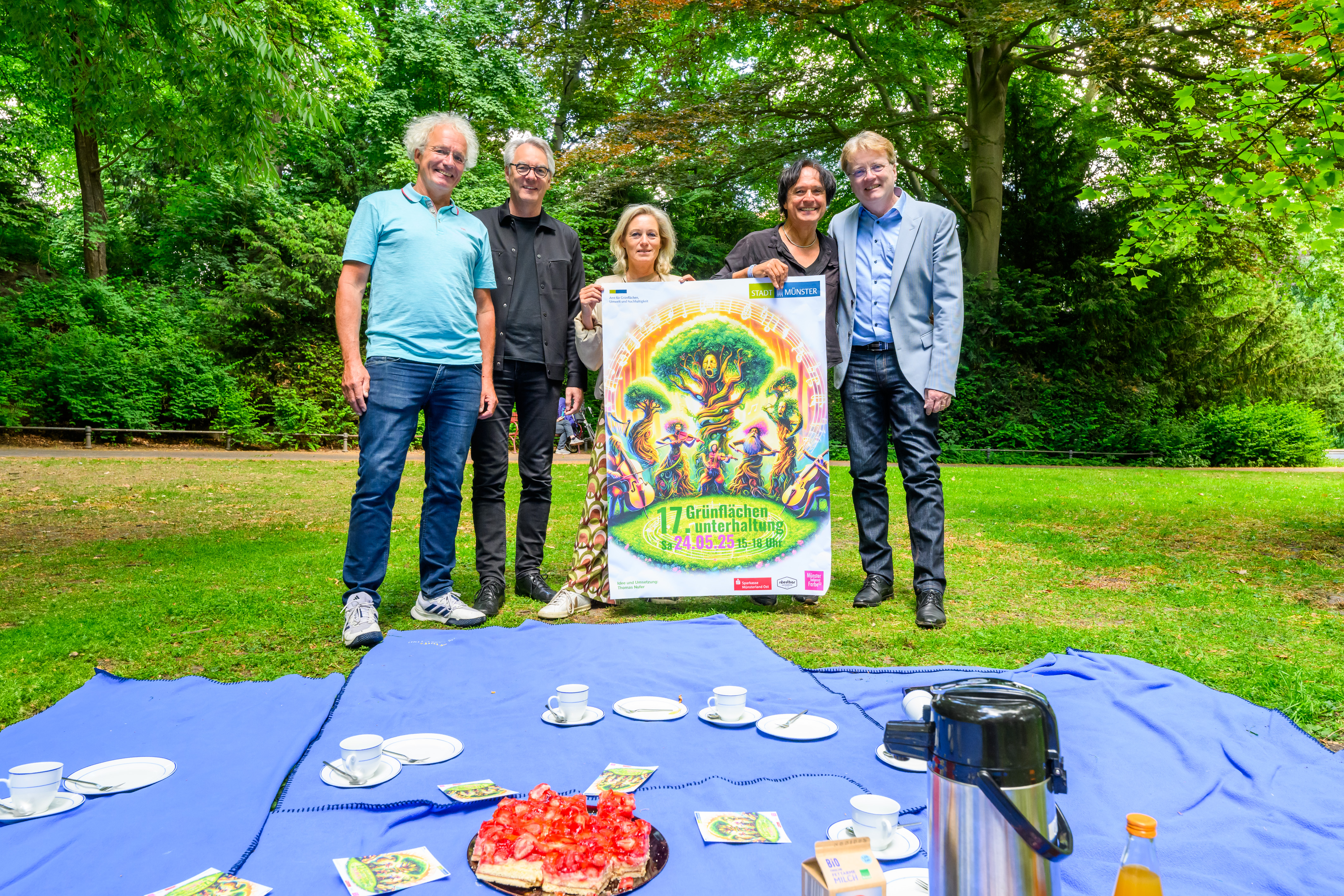 Fünf Personen stehen auf einer Wiese hinter einer Picknickdecke und zeigen ein Plakat