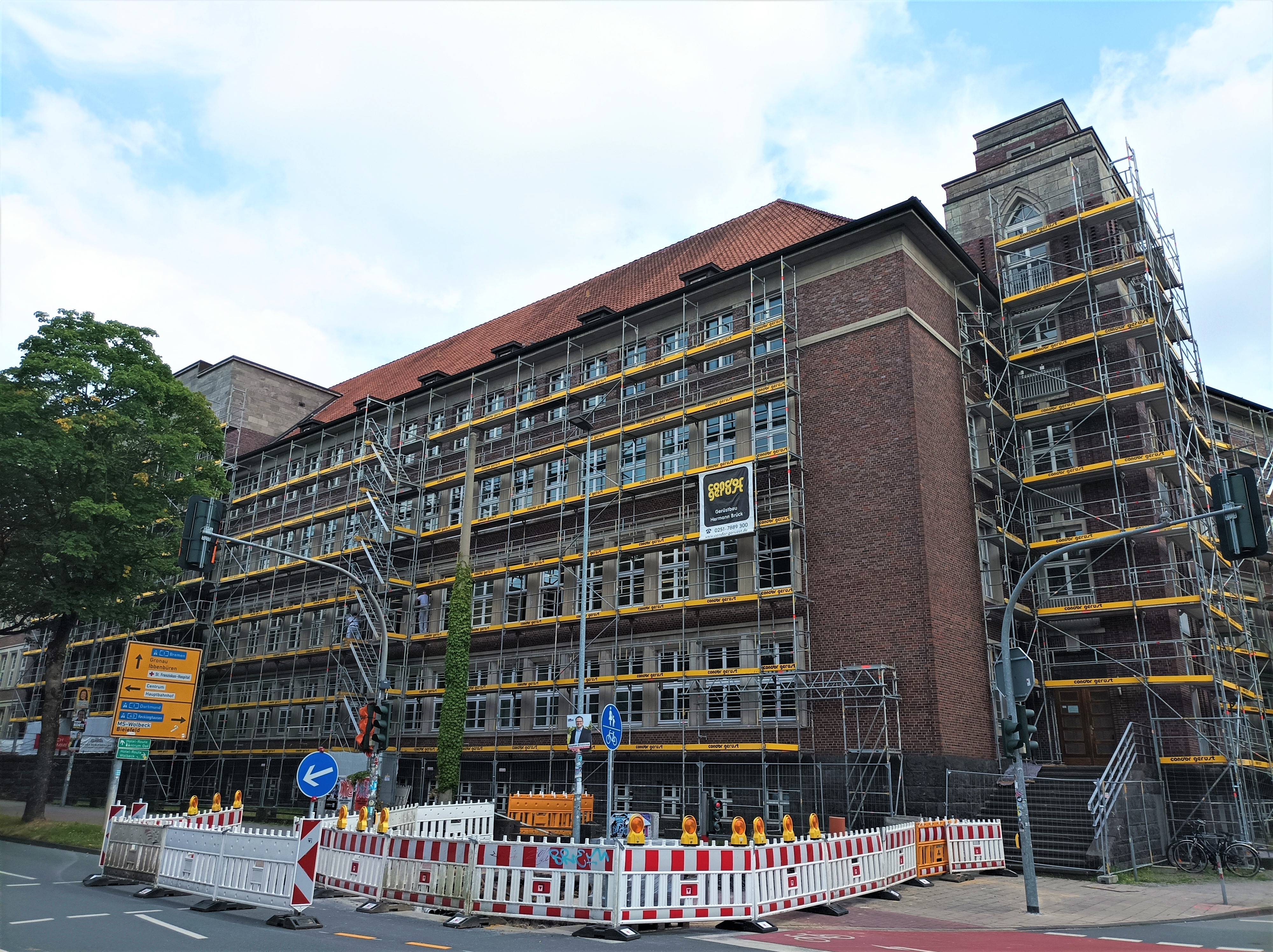 Das Gebäude des Berufskollegs, dessen Fassade mit Baugerüsten verkleidet ist. Es liegt direkt an der Straße und im Vordergrund sind weitere Baustellenabsperrungen, die bis auf die Straße reichen.