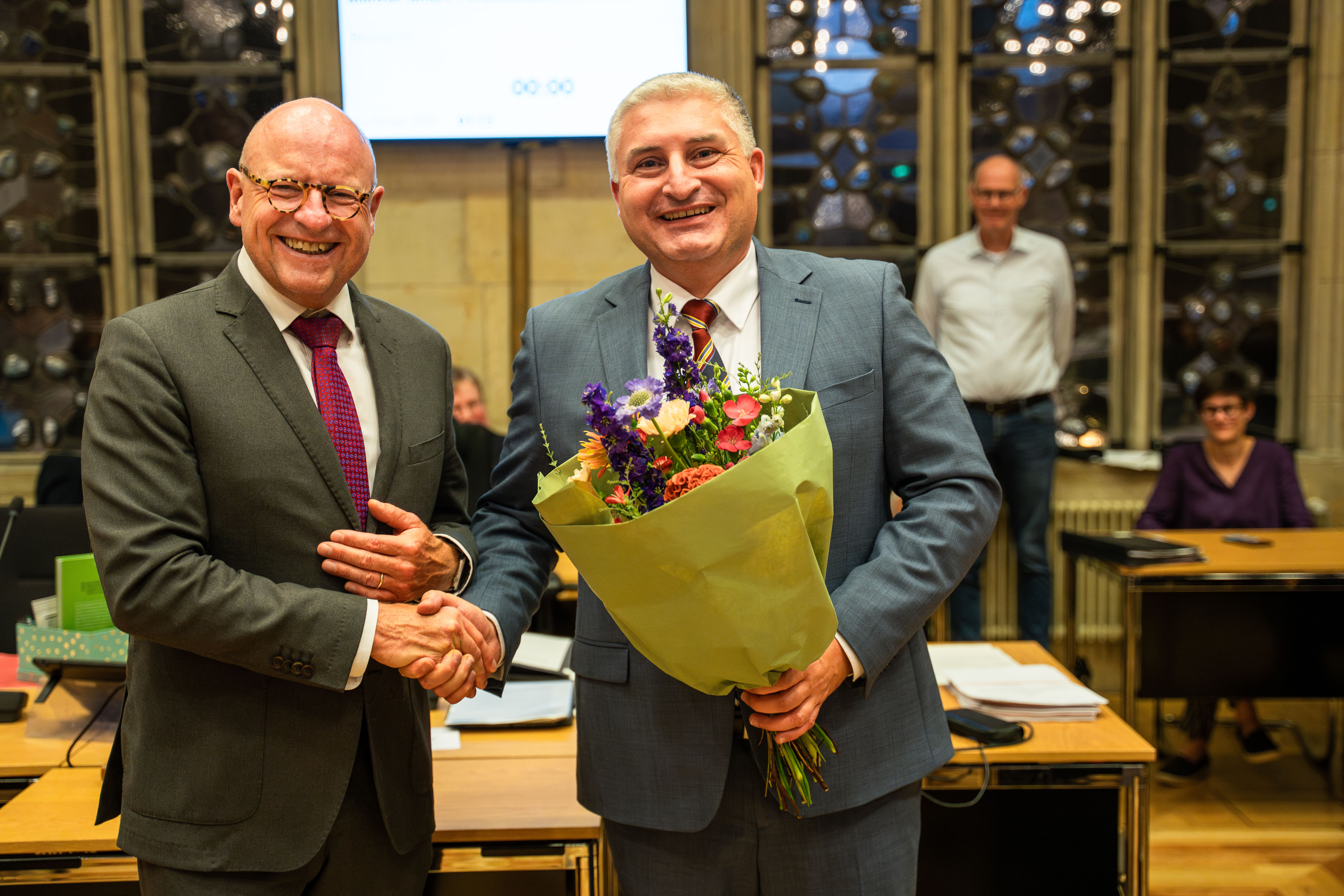 Zwei Männer schütteln sich die Hand; einer hält einen Blumenstrauß in der Hand