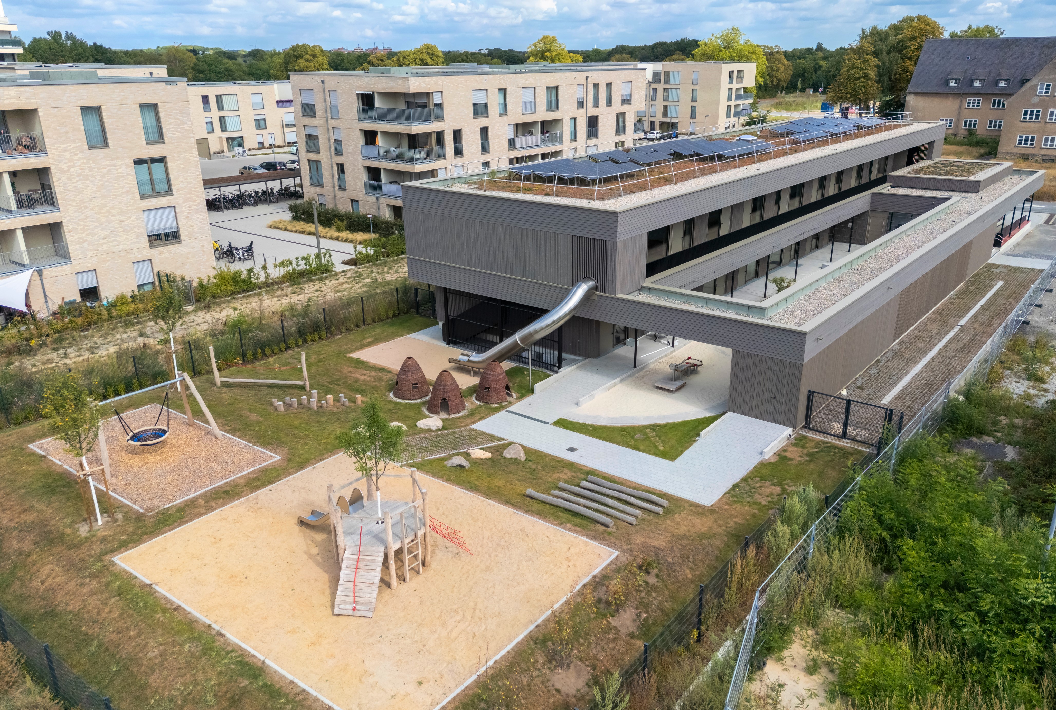 Blick von oben auf ein längliches Gebäude und einen mit Spielgeräten versehenen Außenbereich