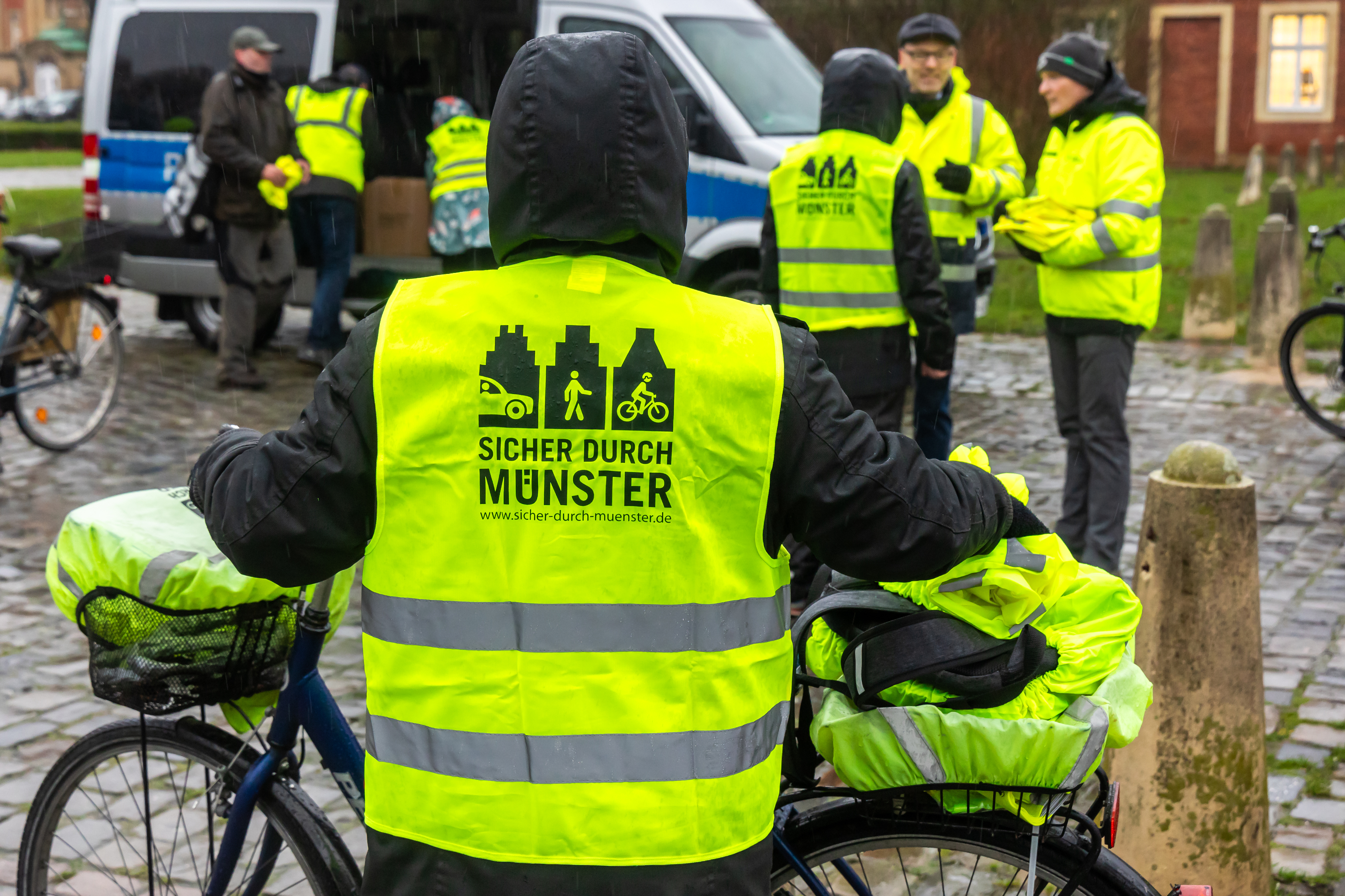 Eine Person steht mit dem Rücken zur Kamera. Sie trägt eine Regenjacke und darüber eine gelbe Warnweste mit der Aufschrift "Sicher durch Münster". 