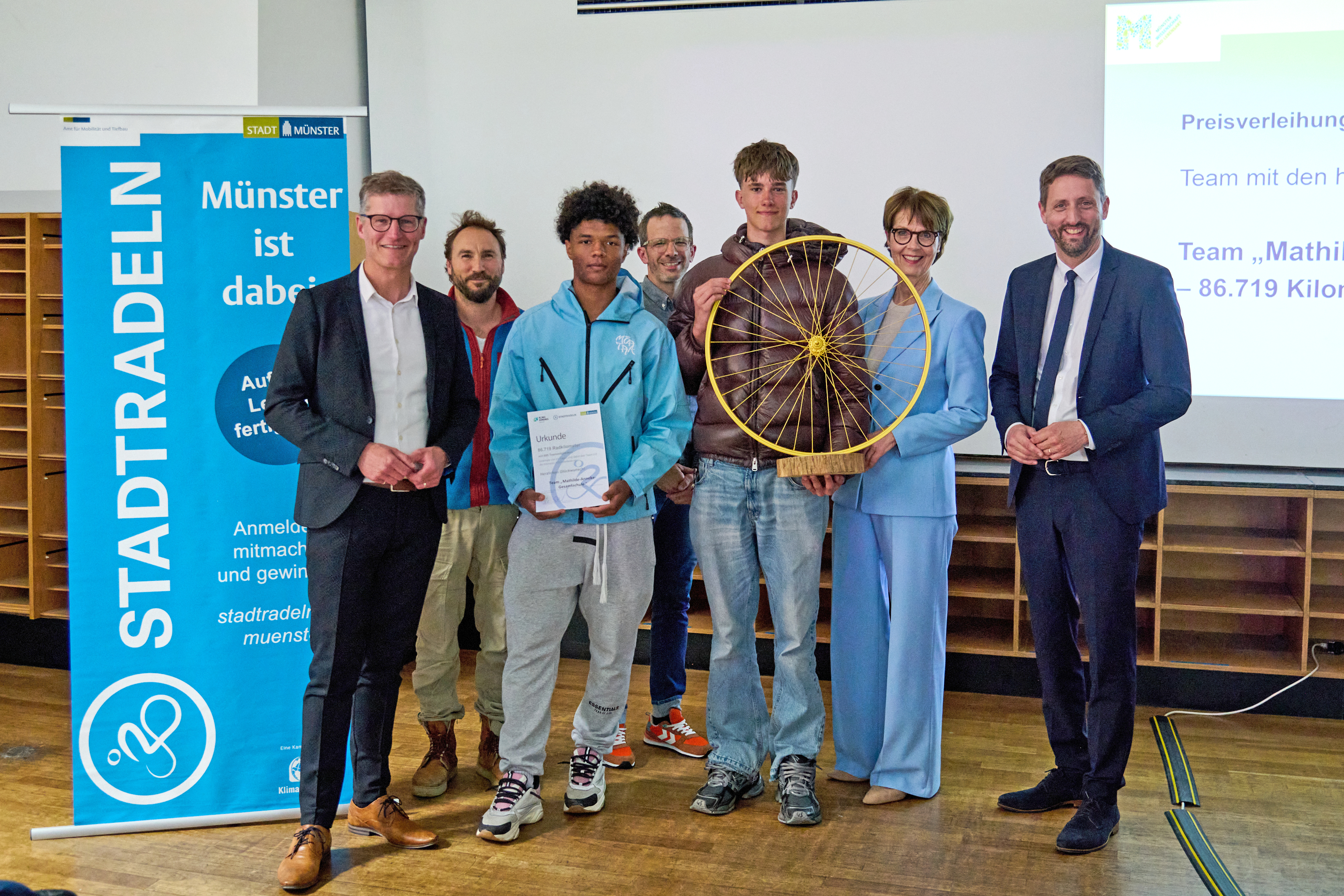 Eine Gruppe von Menschen lächelt in die Kamera, die mittleren zwei halten ein goldenes Rad